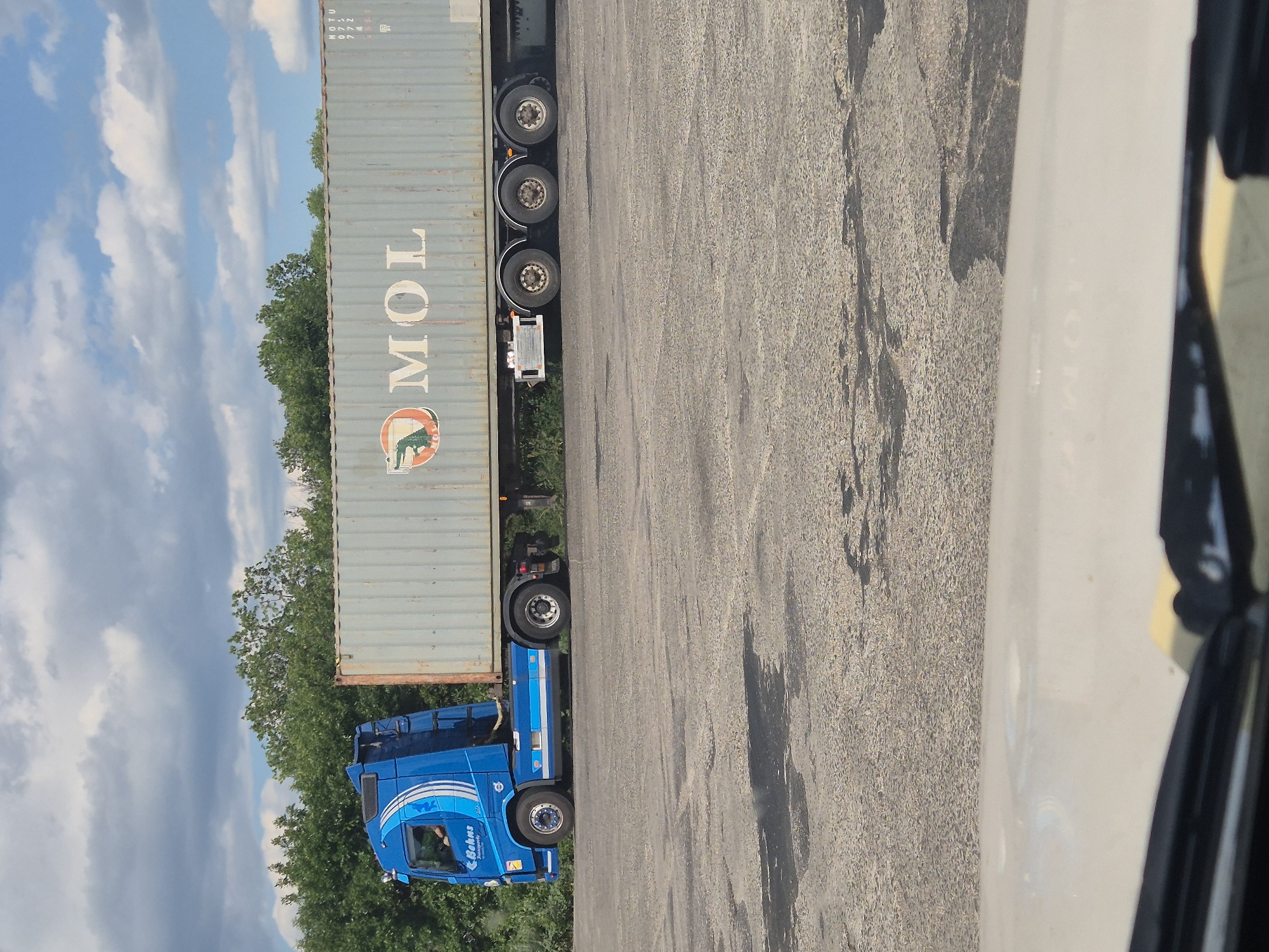 A blue semi-truck with a grey shipping container is parked on an asphalt surface, likely a lot or road, in Pidhaitsi, Ukraine. The truck has the name "Bohns Transport" visible on the side of the cab. The shipping container has the logo and text "MOL" printed on it. Green trees are visible in the background, and a cloudy blue sky is overhead. The asphalt surface shows signs of wear and tear, with some dark patches possibly indicating oil stains. The image appears to be taken from inside another vehicle, as the bottom of the frame shows a portion of a car's interior, including the windshield wiper and dashboard.