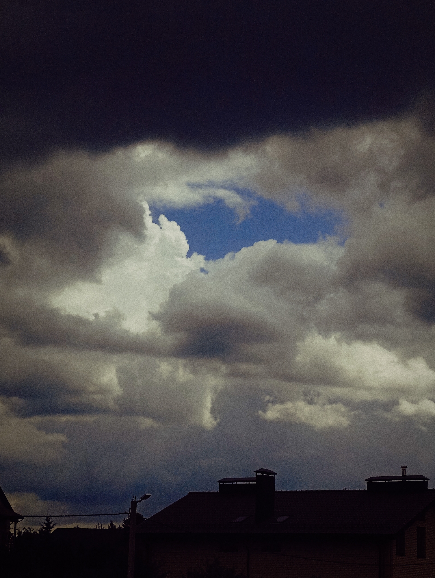 The image captures a dark and cloudy sky over a residential area. The clouds are heavy and varied in shade, ranging from deep gray to white, with a patch of blue sky visible in the center. In the foreground, the silhouettes of rooftops with chimneys are visible, indicating houses. A street lamp and some trees are also discernible in the lower part of the image. The overall atmosphere is somber and moody, suggesting overcast weather, possibly before or after a storm. The location is identified as Unknown, Unknown.