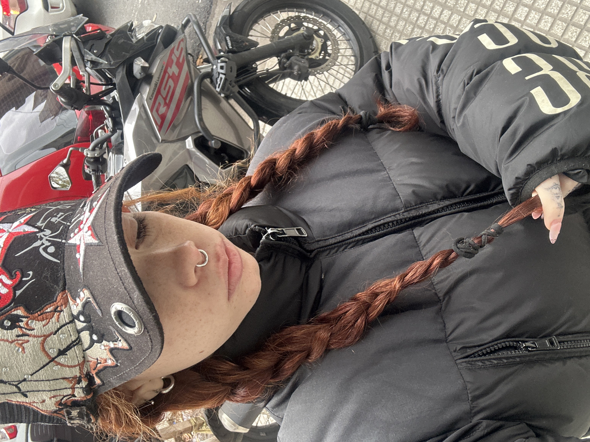 A person is lying on the ground next to a motorcycle. The person is wearing a black puffer jacket with white lettering, a black baseball cap with an embroidered design, and has red hair in braids. The motorcycle is red and gray and appears to be parked on a sidewalk with patterned tiles. The location is presumed to be Buenos Aires, Argentina.