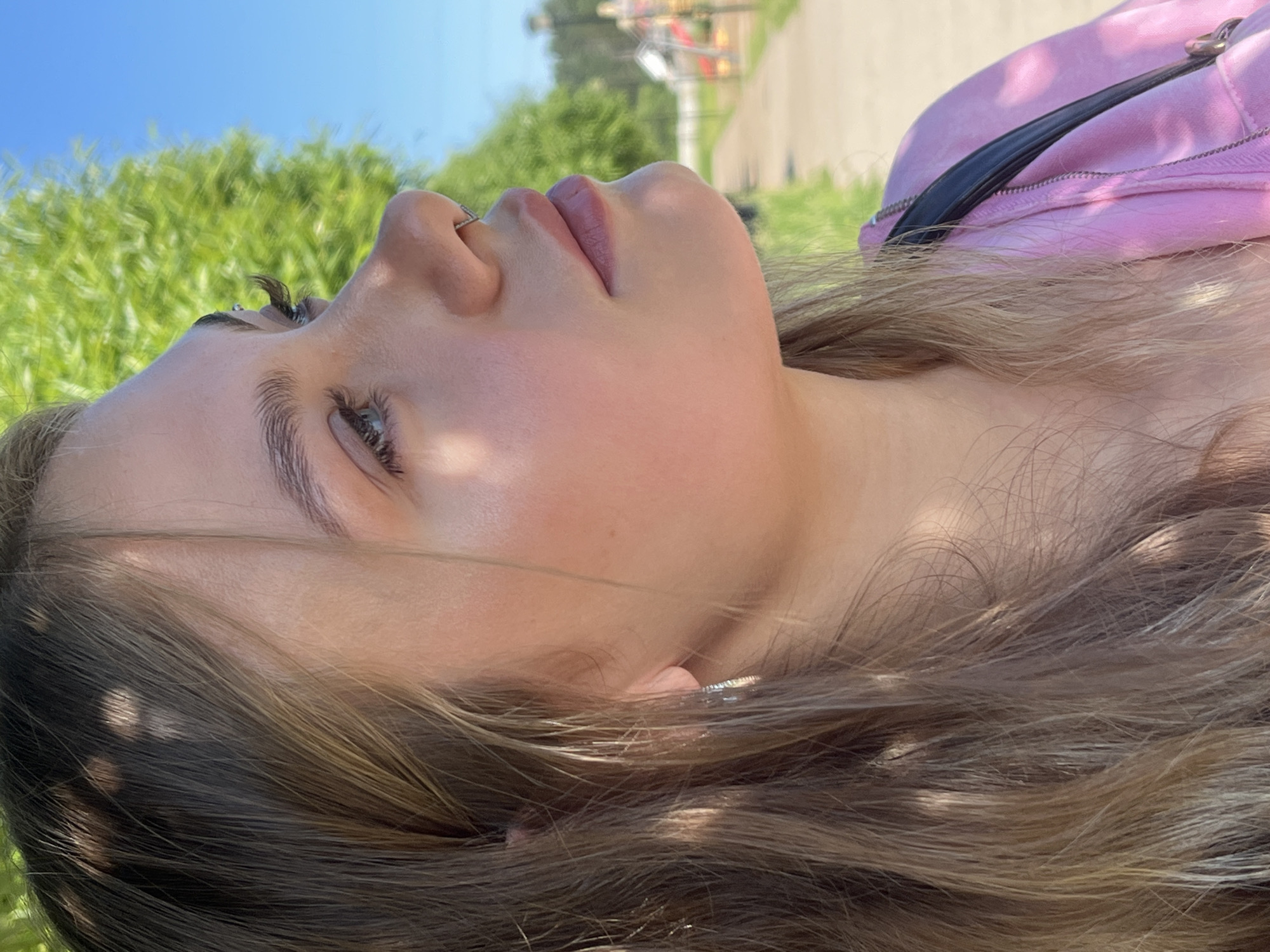 This is a close-up, profile shot of a young woman with long, light brown hair, wearing a pink hooded jacket and a black strap across her shoulder. She has natural makeup, including eyeliner and mascara, and a nose ring. She is outdoors with a bright blue sky and lush green trees in the background, suggesting a sunny day. The scene is likely a park or a similar outdoor public space in Chaikovsky, Russia, given the presence of a pathway and distant playground equipment. The lighting indicates it's daytime, and the dappled light on her hair suggests she is positioned under a tree or similar foliage.