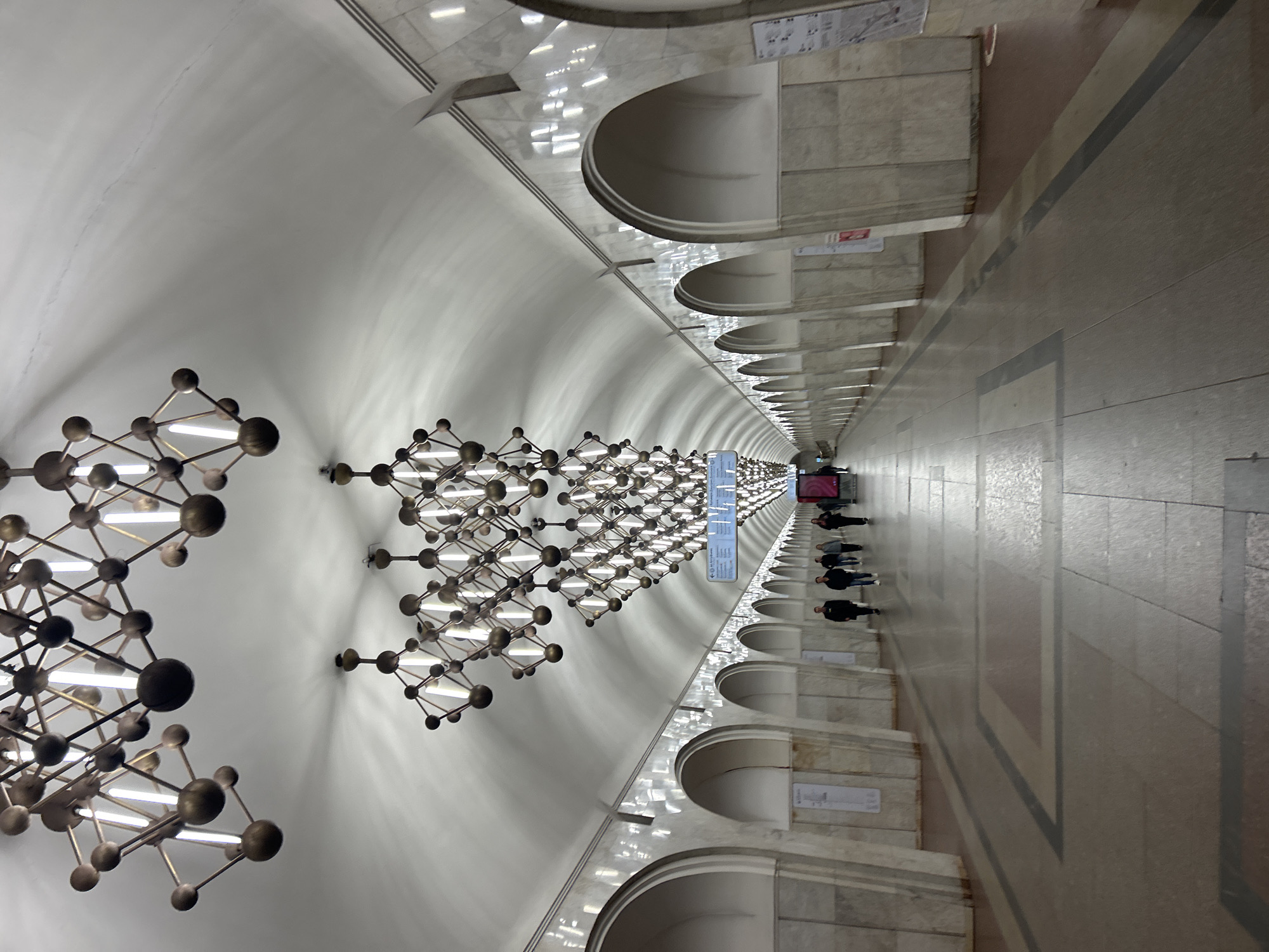 A subway station in an unknown city features a long, arched ceiling with distinctive light fixtures resembling molecule models. People are walking along the platform. The walls are marble with arched recesses, and signage is visible. The floor is patterned with geometric shapes.