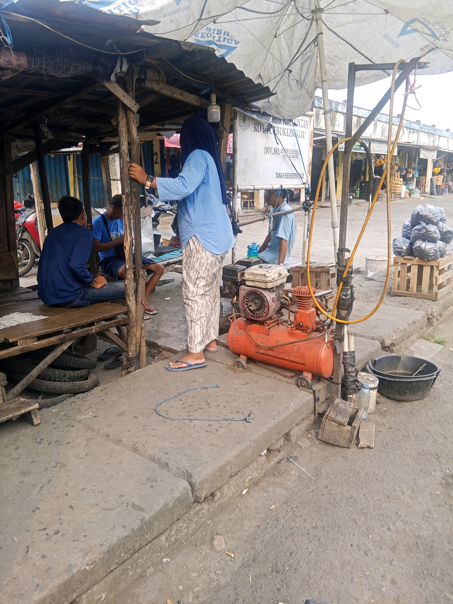 The image depicts an outdoor scene in what appears to be a market or roadside stall area in Lubuklinggau, Indonesia. In the foreground, a person wearing a blue long-sleeved shirt and a headscarf stands with their back to the camera, facing an orange air compressor. They are wearing patterned pants and flip-flops. To the left, two men are seated on a wooden bench under a makeshift shelter. One man, wearing a blue long-sleeved shirt and jeans, is looking down. The other man, in a blue t-shirt and dark shorts, is also looking down and appears to be holding something. The shelter is constructed with rough wooden poles and a tattered canvas roof.
In the middle ground, the orange air compressor is connected to yellow hoses that are wrapped around a wooden pole. Beside the compressor is a black plastic tub. Behind the compressor, a wooden crate is stacked with several black plastic bags. The ground is paved with concrete slabs and asphalt. Further in the background, a row of stalls or shops can be seen, with various items displayed. A sign with text in Indonesian is visible on a white board, reading "PEMERINTAH KOTA LUBUKLINGGAU". Motorcycles are parked to the left of the scene. The overall environment suggests a busy, functional outdoor commercial space.