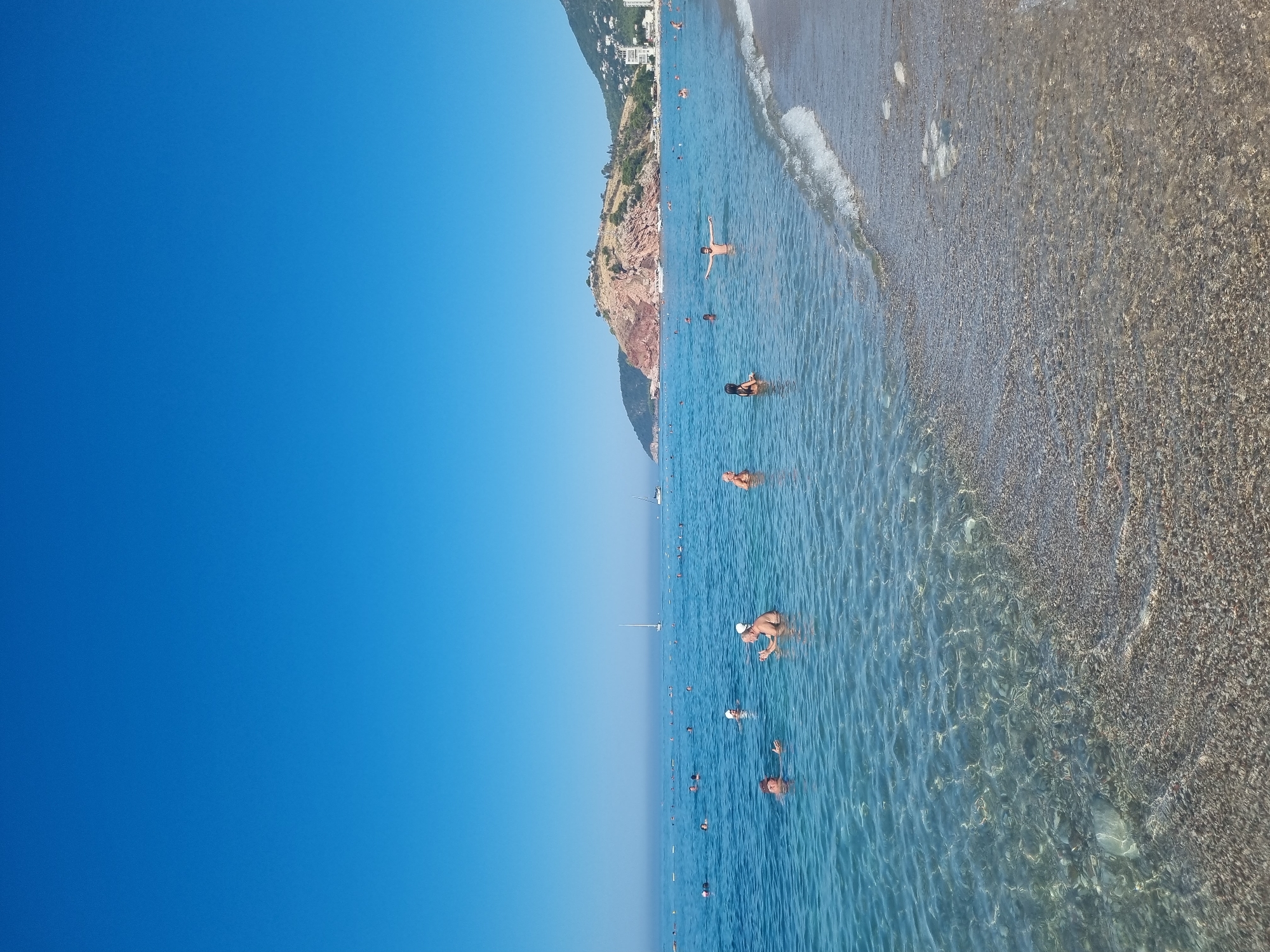 The image captures a sunlit beach scene in Sutomore, Montenegro, with the Adriatic Sea stretching towards the horizon under a clear blue sky. The water is clear and shallow near the shore, revealing a pebble-strewn seabed. Several people are visible in the water, some swimming and others standing or interacting. In the foreground, two adults are in the water, appearing to converse. Further out, a young person stands with arms outstretched. In the mid-ground, more individuals are dispersed throughout the water, enjoying the sea. A few sailboats are dotted on the distant water. Along the coastline in the background, a rocky hill covered in vegetation rises prominently, with buildings clustered on the slopes of another hill to its right, indicating the presence of the town. The beach itself is a mix of sand and pebbles.