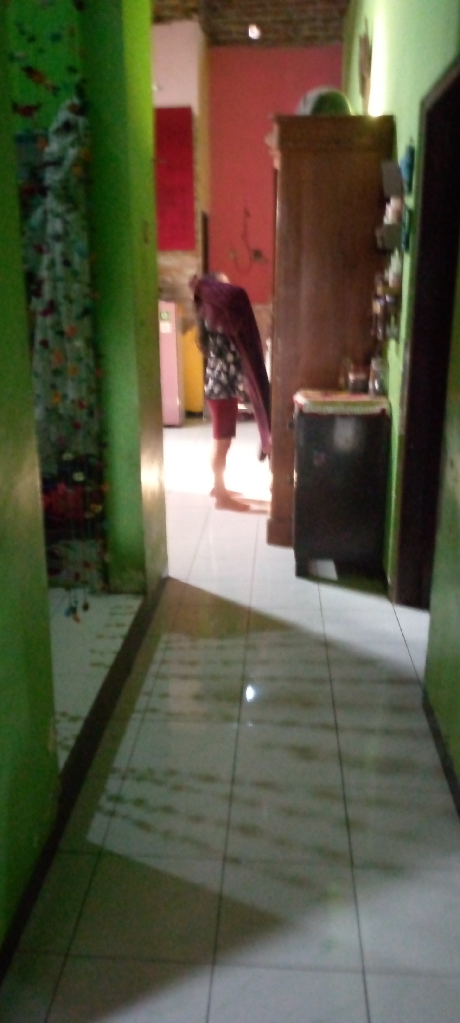A low-angle, blurred, full shot captures an indoor hallway scene. On the left, a green wall is visible, partially obscured by a patterned curtain. Directly ahead, a person is walking away from the camera, draped in a dark reddish-brown cloth. They are wearing a patterned top and red shorts, with bare feet. To the right of the person, a wooden cabinet stands next to a dark object, possibly a trash bin. In the background, a pink wall and a yellow object are visible. The floor is tiled with light-colored squares, with shadows cast across it. The lighting suggests it is daytime, with sunlight entering from the right.