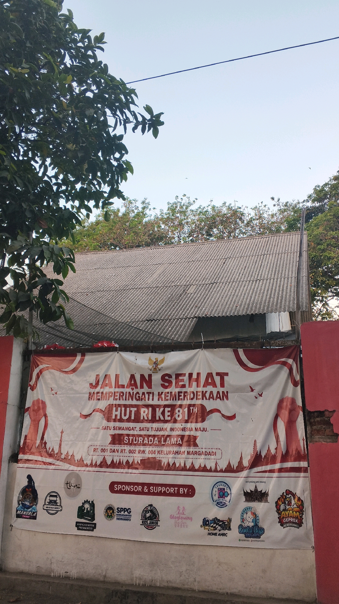A banner displays "JALAN SEHAT MEMPERINGATI KEMERDEKAAN HUT RI KE 81TH" meaning "Healthy walk commemorating the 81st Independence Day of the Republic of Indonesia." Below this, it states "SATU SEMANGAT, SATU TUJUAN, INDONESIA MAJU." which translates to "One Spirit, One Goal, Advanced Indonesia." The banner also includes "STURADA LAMA RT. 001 DAN RT. 002 RW. 004 KELURAHAN MARGADADI" indicating the location as Sturada Lama, neighborhood RT 001 and RT 002, RW 004, Margadadi Village in Indramayu, Indonesia. Various sponsor logos are displayed at the bottom of the banner. The banner is hung on a corrugated roof structure with red painted walls on the sides. Trees with green leaves are visible in the background, along with a clear sky and a power line.