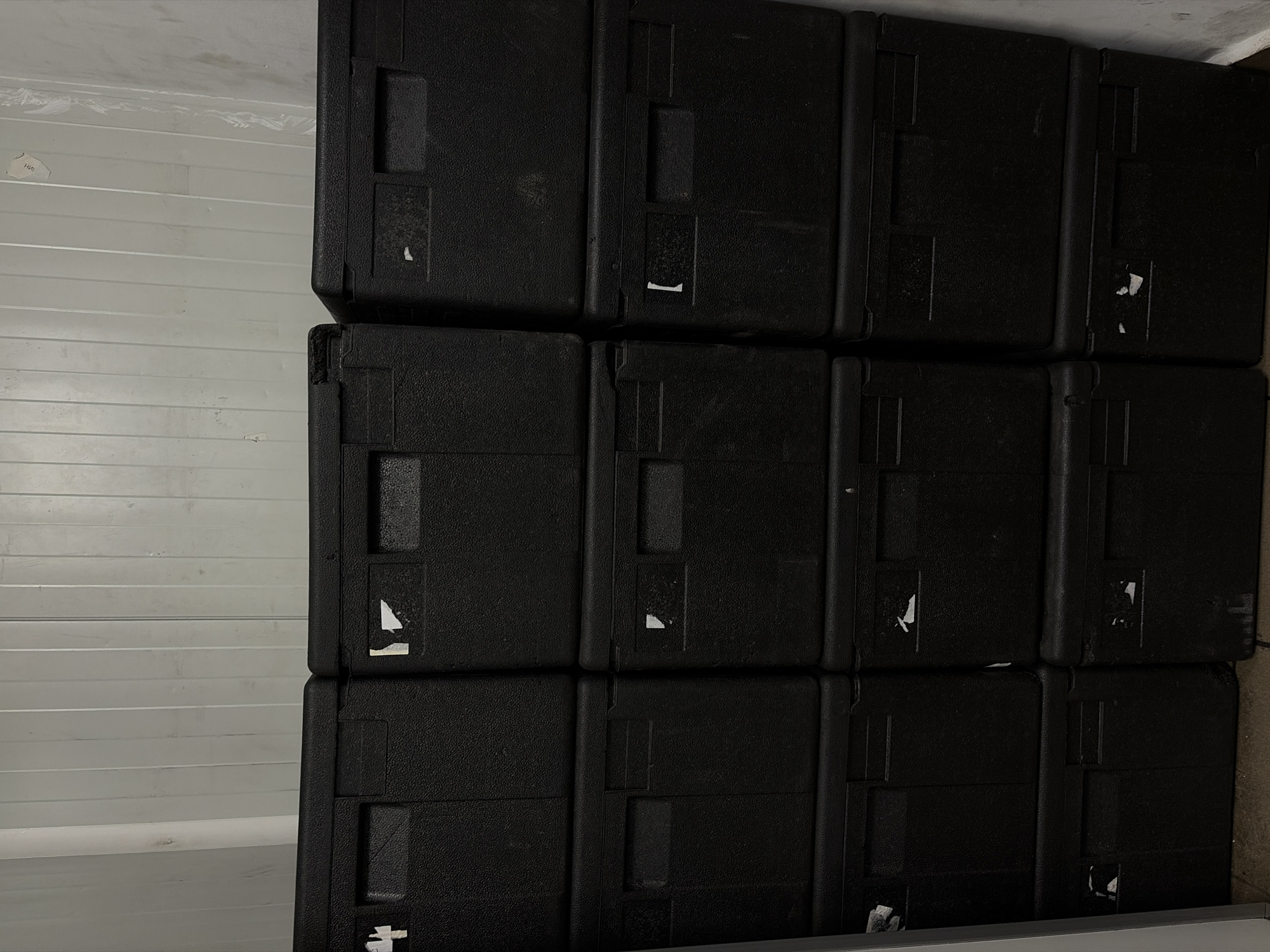 A stack of black, rectangular cases is positioned against a wall with white horizontal panels. The cases are arranged in three rows and four columns. Some of the cases have small, rectangular indentations or handles. Small, white fragments, possibly remnants of stickers or tape, are visible on several cases. The location is an indoor space in Sofia, Bulgaria.