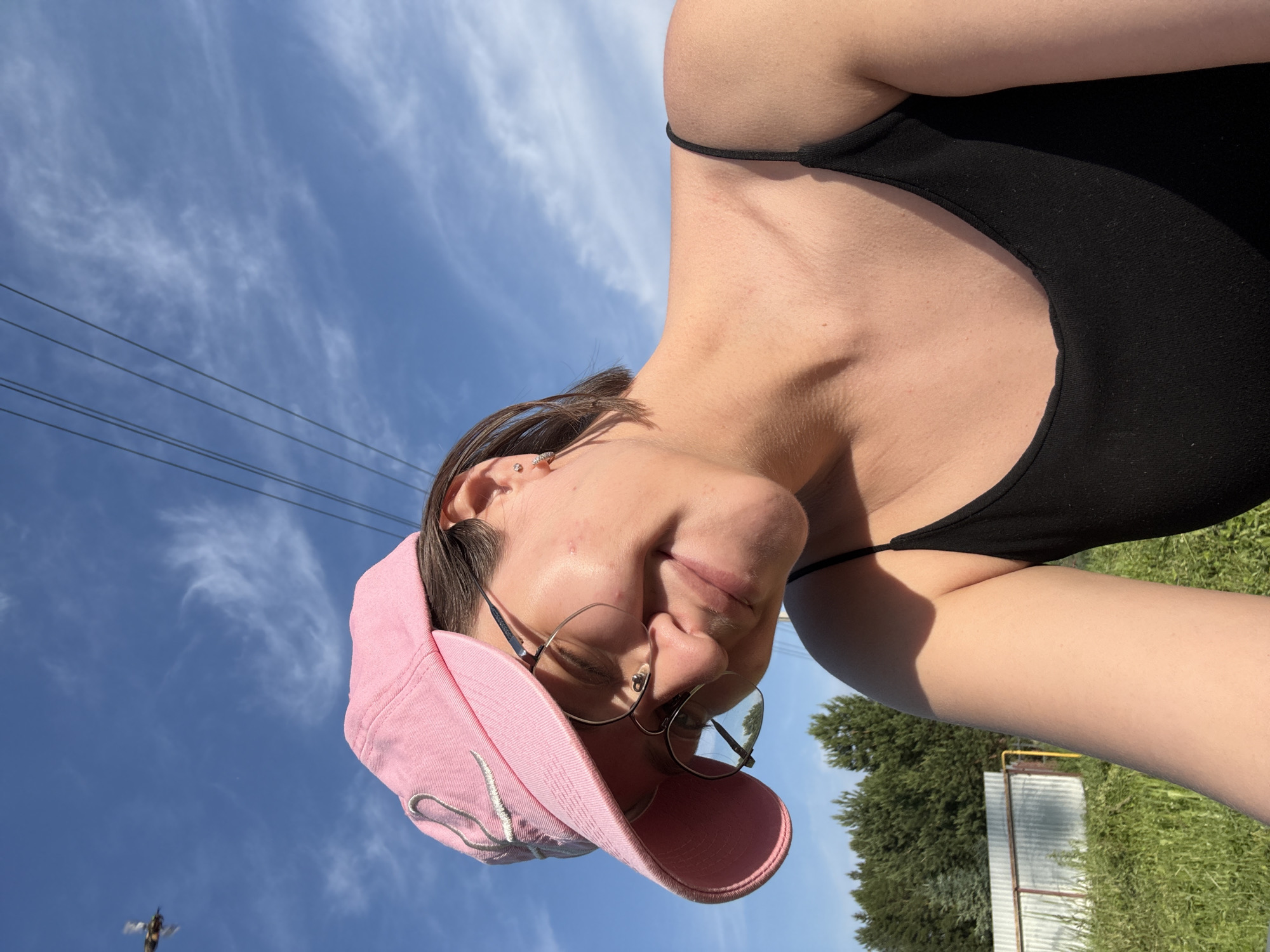 A young person with short brown hair, wearing a pink baseball cap and eyeglasses, looks down with a slight smile. They are wearing a black tank top. The background shows a bright blue sky with wispy clouds and power lines. Below the sky, there are green trees and a fence. In the distance, a small flying object is visible against the sky. The scene appears to be outdoors, possibly in a rural or suburban area.