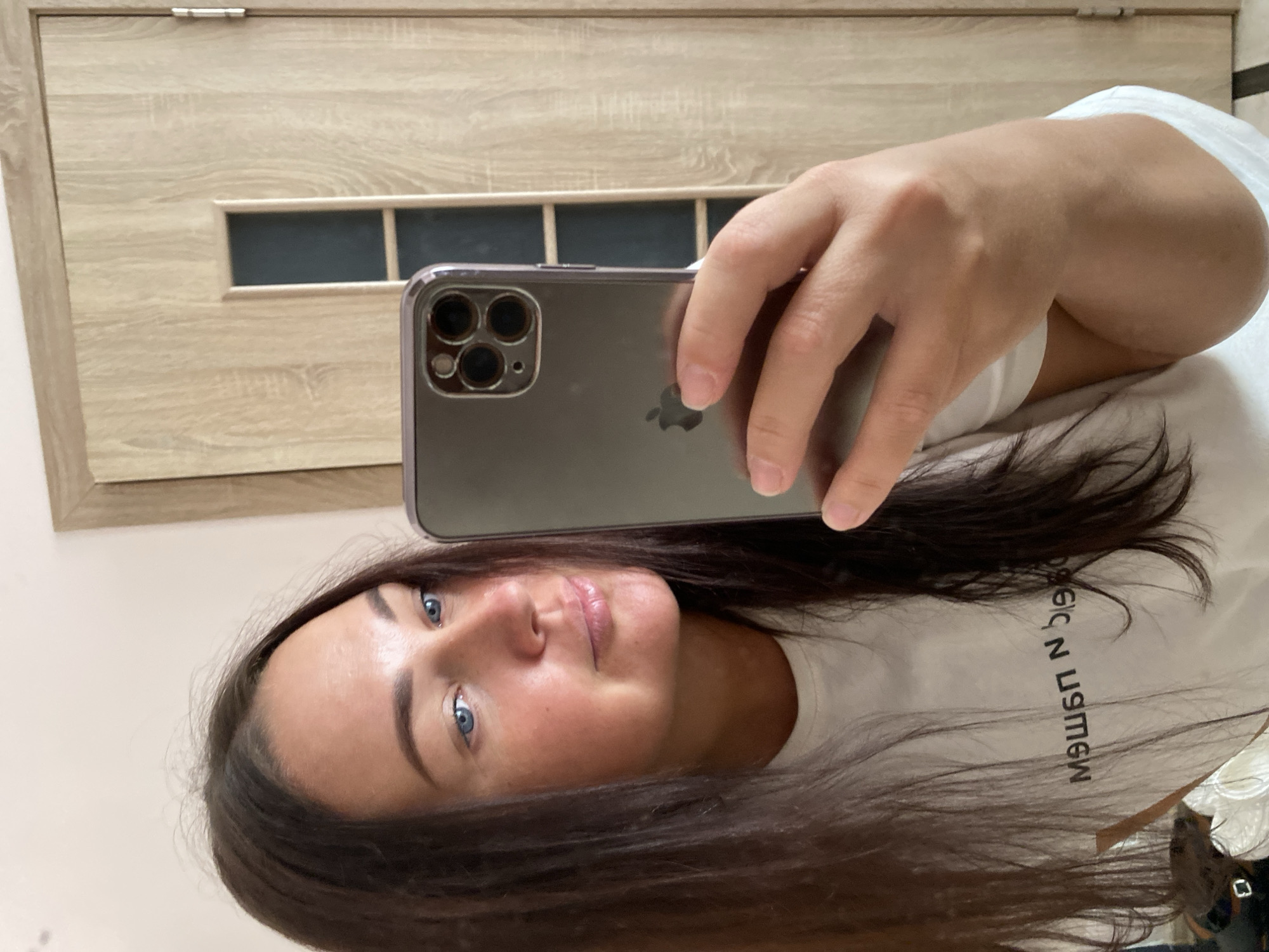 A woman is taking a mirror selfie in a room. She has long, dark hair, blue eyes, and is wearing a white t-shirt with text printed on it. She is holding a silver smartphone in her right hand. The background shows a wooden door with a frosted glass panel and a light-colored wall. The room appears to be indoors, likely a residential setting. There is no indication of the city of Sochi, Russia.