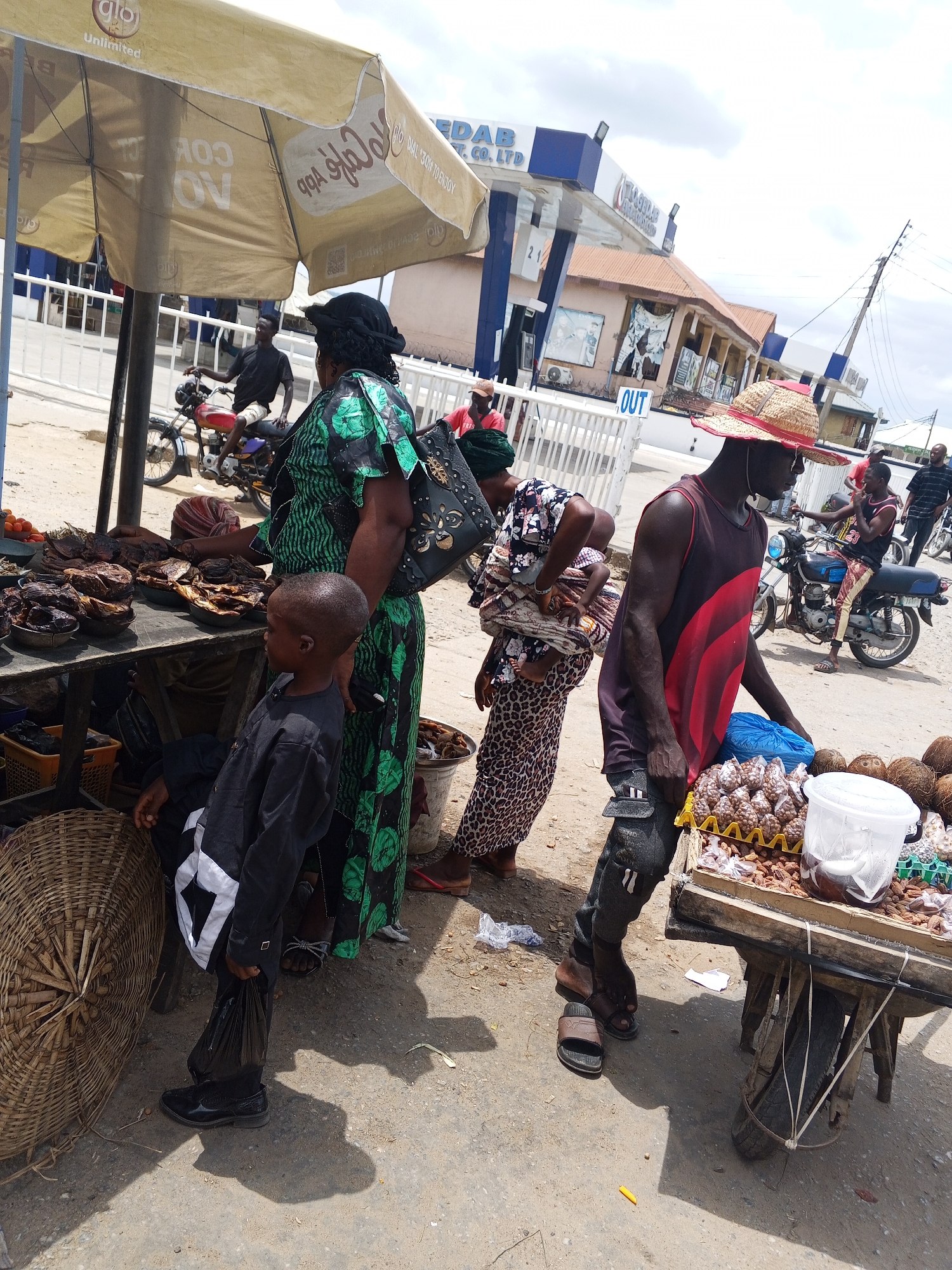 In Zango-ndaji, Nigeria, an outdoor street scene shows several vendors and customers at makeshift stalls on a bright day. One vendor, wearing a straw hat and a red and black tank top, stands beside a wooden cart laden with nuts and coconuts. A woman in a green patterned dress and a child in a black outfit are at a stall displaying smoked fish under a large umbrella. Another woman carries a child wrapped in fabric. In the background, people are on motorcycles and on foot near a building marked with "OUT."