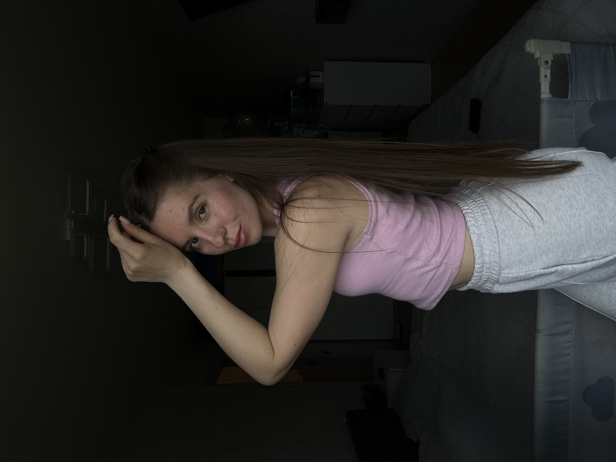 A young woman with long, brown hair is lying on a bed. She is wearing a pink tank top and gray sweatpants. She is resting her head on her hand and looking at the camera. The background is dark, but some furniture is visible, including a white dresser and a crib. The location may be Klin, Russia, based on the prompt.