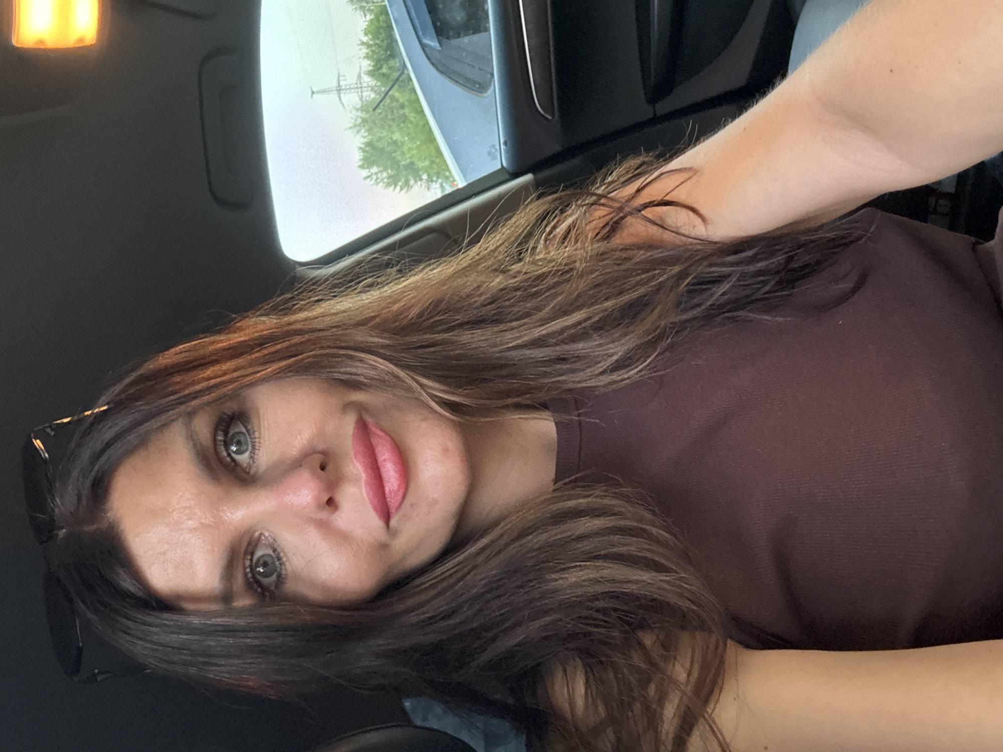 A woman with long, dark brown hair is photographed from the chest up, looking directly at the camera. She is sitting in the driver's seat of a car, with sunglasses perched on top of her head. Her expression is serious, with her eyes wide open. She is wearing a dark brown, sleeveless top. The interior of the car is visible, including part of the dashboard and the roof with an illuminated light fixture. Outside the car window, trees and power lines can be seen against a pale sky, suggesting an urban or suburban environment. There is another vehicle visible through the window behind her. The lighting suggests it is dusk or early evening.