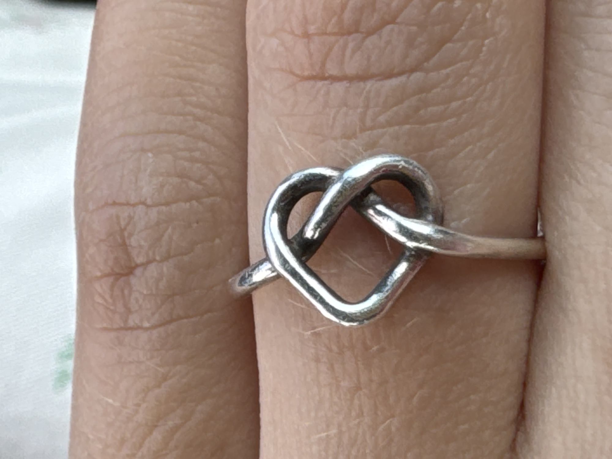 A close-up image shows a person's finger with a silver ring in the shape of a heart-shaped knot. The ring is positioned on the middle finger. The finger shows skin texture details. The background includes a blurred white surface. The location is unknown.