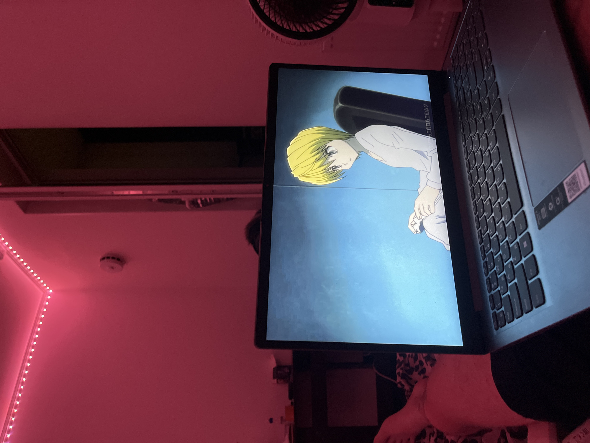 A laptop is positioned in a room illuminated by pink LED strip lights. An anime character with blonde hair is visible on the laptop screen. A portable fan is seen behind the laptop. A partial view of a person's legs is present in the foreground.