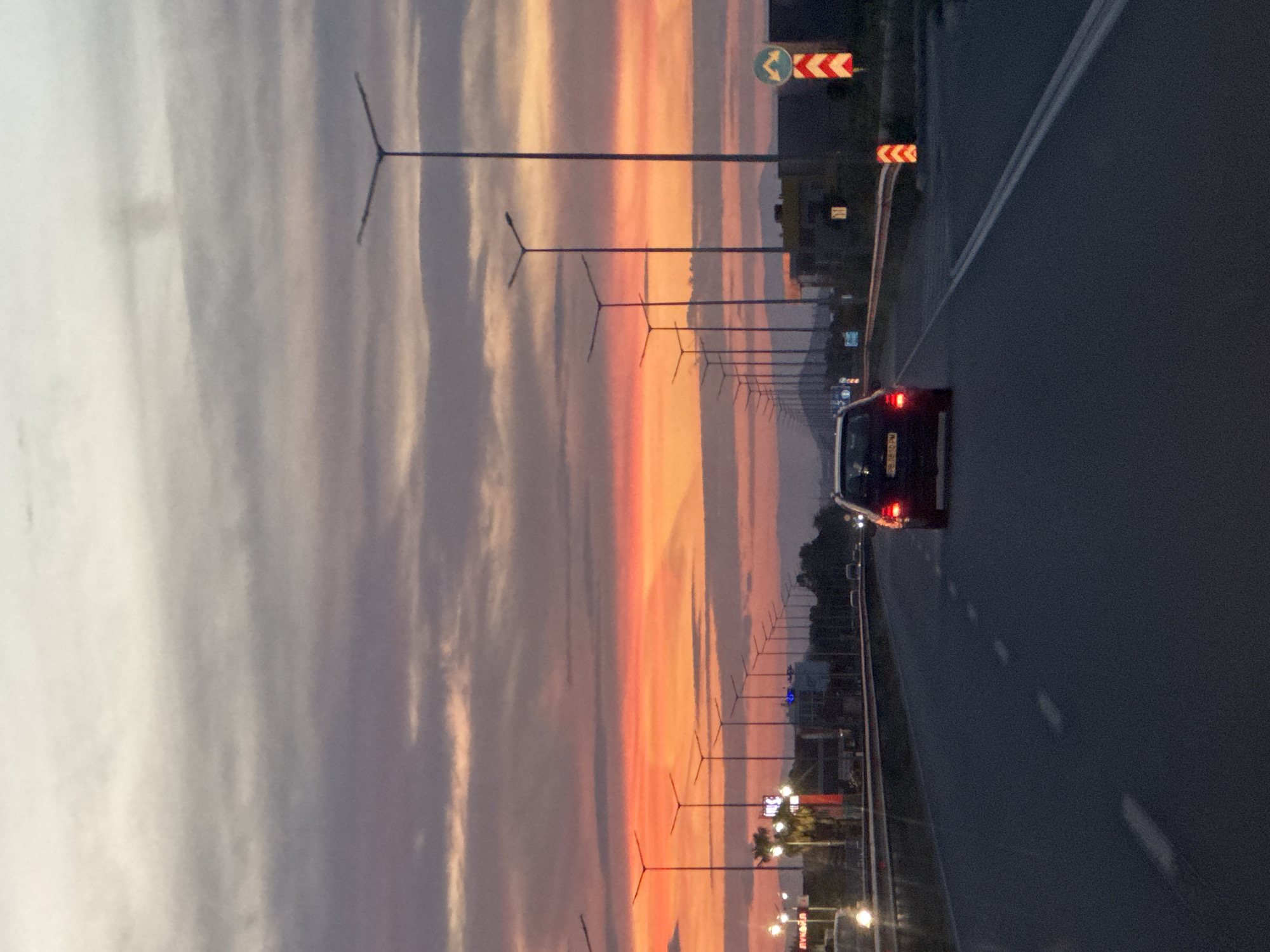 The image shows a road at sunset in Asenovgrad, Bulgaria. A car with its headlights on is traveling along the road. Several streetlights are visible along the side of the road. The sky is filled with colorful clouds in shades of orange, pink, and gray. Road signs are present. Buildings and trees can be seen in the background.