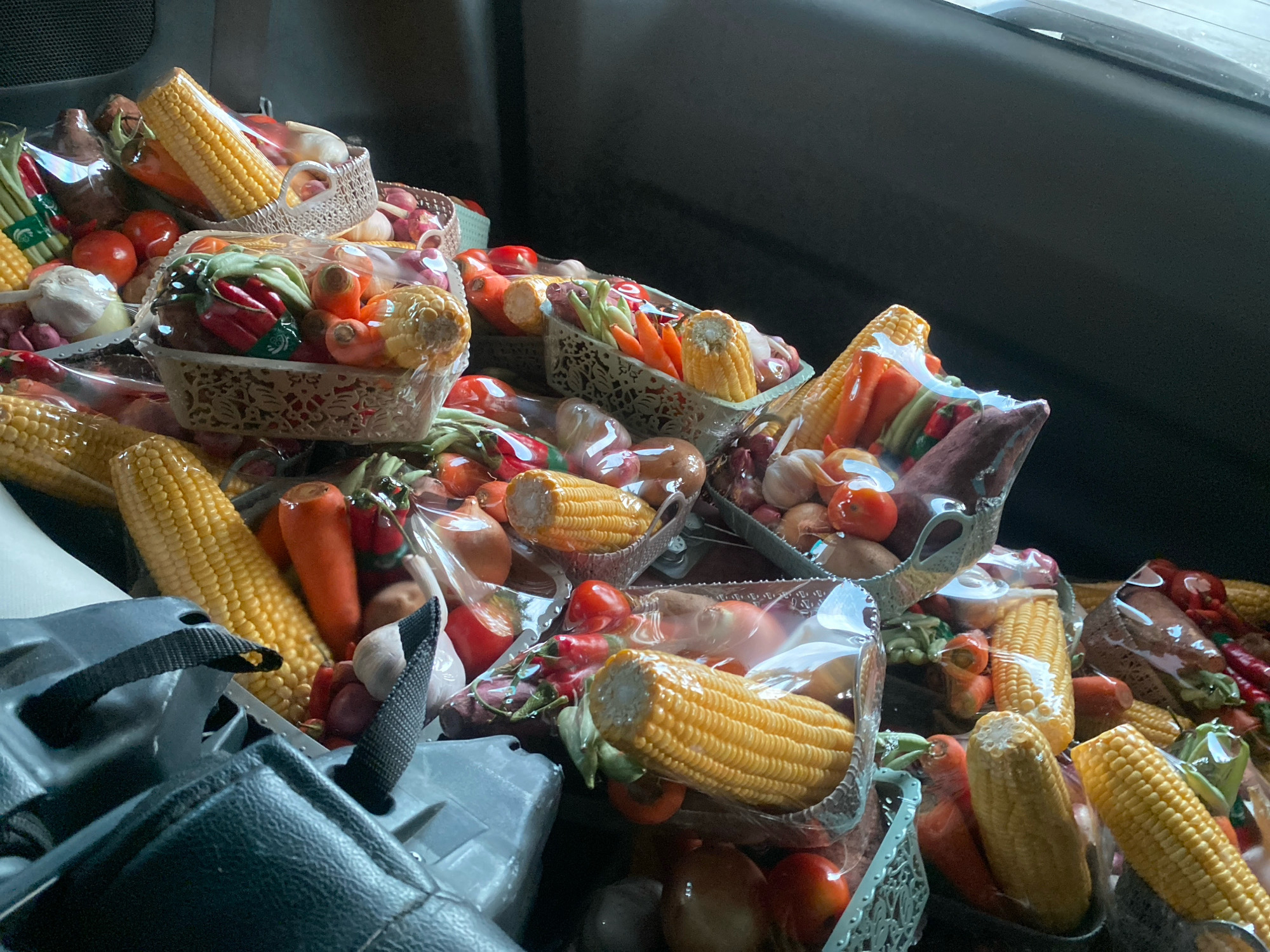 In a vehicle, likely a car, in Banjarbaru, Indonesia, numerous baskets filled with fresh produce are piled up. The baskets contain various vegetables and fruits such as corn on the cob, carrots, tomatoes, onions, chili peppers, and garlic. Many of these items are individually wrapped in clear plastic, suggesting they are prepared for sale or distribution. The overall scene depicts a substantial quantity of fresh food packed tightly within the confines of the vehicle's interior, possibly the back seats or trunk area. The lighting is somewhat dim, indicating an indoor setting or possibly the interior of the vehicle with limited external light.