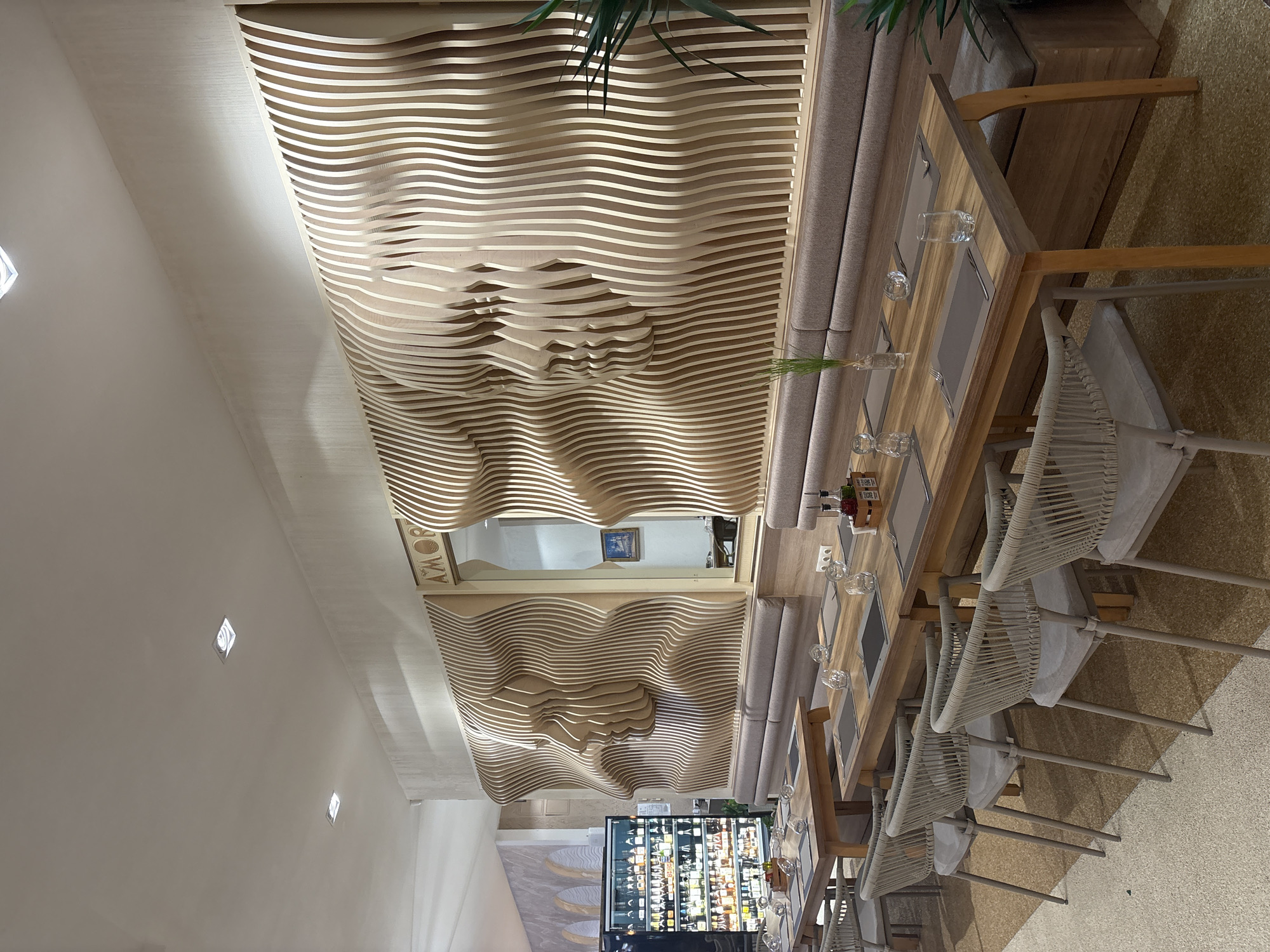 The image shows an interior view of a restaurant. The dominant feature is a large, abstract wooden art installation on the wall, depicting layered profiles of faces. The installation is illuminated from behind, creating a shadow effect. Below the art installation, there is a long, cushioned banquette seat. In front of the banquette, several wooden dining tables are set with placemats, cutlery, glassware, and small decorative items like a vase with a plant and bottles of condiments. Chairs with woven backs and cushioned seats are placed around the tables. To the left of the art installation, there is a refrigerated display case filled with bottles of alcohol, suggesting a bar area. The overall lighting in the restaurant is soft and ambient. The floor has a textured, light-colored finish. The image is a static shot taken from a slightly elevated angle, looking across the dining area towards the art installation.