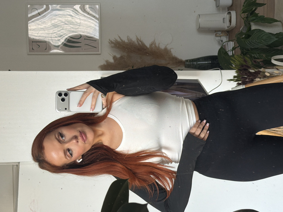 An adult female is depicted in a mirror selfie taken indoors. She has long, reddish-brown hair, visible makeup, and a white wireless earbud in her left ear. She wears a white ribbed sleeveless crop top, black long-sleeved arm warmers with thumbholes, and black ribbed high-waisted leggings. A ring with a dark-colored stone is present on her right ring finger. She holds a light-colored smartphone with a triple-lens camera in her right hand, oriented to capture her reflection. Her left hand rests on her hip. The background, visible in the mirror, features plain white walls. A framed abstract art print with undulating patterns and partially visible text hangs on the wall behind her right shoulder. Below it, a tall, dark green vase contains dried pampas grass. Further to the right, a white pet feeder with two bowls is visible on a surface, alongside other potted plants and decorative objects. A large, dark-leafed plant is partially visible on the left side of the frame. The precise location within the city of Paris, France, cannot be determined from the visual evidence.
