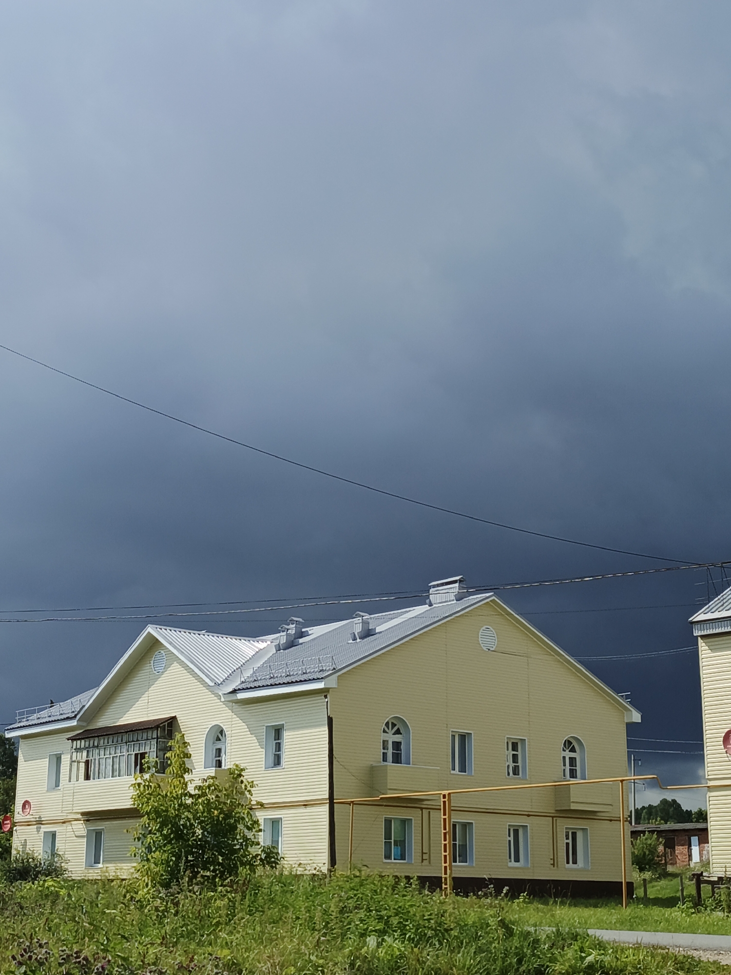 This image shows a light yellow, two-story building with a silver roof, located in Medvedyovka, Russia. The building has a small balcony with windows on the second floor, and multiple rectangular windows on both floors. A cluster of green bushes and grass is in the foreground. The sky is overcast with dark, heavy clouds. Power lines are visible against the sky. The scene appears to be a residential area.