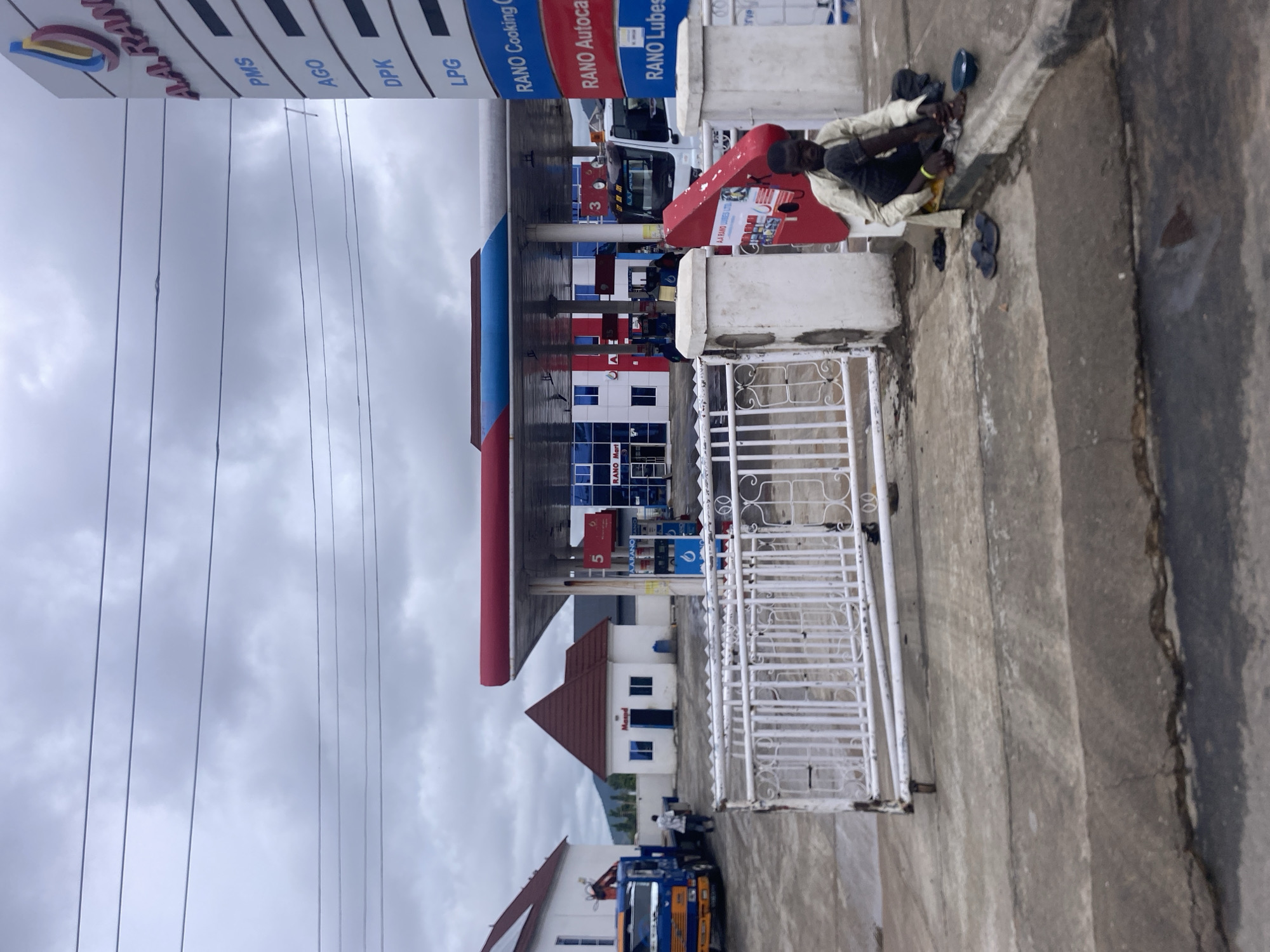 The image shows a RANO filling station located in Jalingo, Nigeria. In the foreground, a man dressed in a light-colored shirt and dark pants is seated on the ground next to a curb, with a small blue bowl in front of him. Several pairs of flip-flops are scattered around him. A white metal gate is positioned behind him. A red and white kiosk-like structure is to his left, with some printed material visible. The filling station features a red and blue canopy over fuel pumps. A white truck with an orange crane is parked to the left of the frame, near a small building with a red tiled roof. Another white truck is visible under the filling station canopy. The sky is overcast.