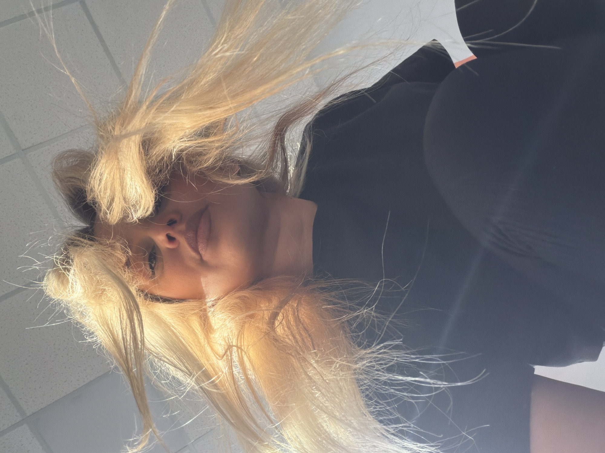 The image is an upward-facing perspective of a woman with long, blonde hair. She is wearing a black shirt and is positioned against a white, tiled ceiling, typical of an office or commercial space. No specific details indicate the location is Sarapul, Russia.
