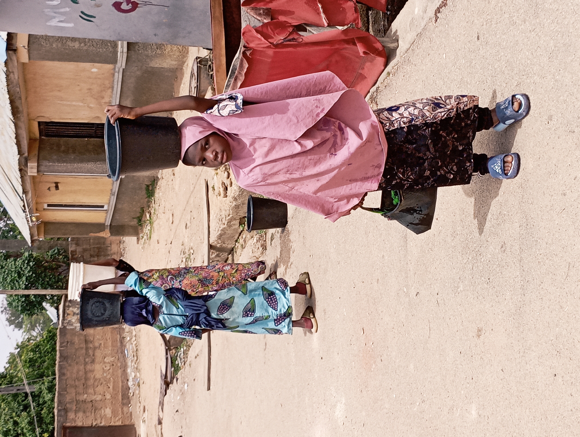 In Jos, Nigeria, two young girls carry water in buckets on their heads down a dusty concrete path beside buildings. The girl in the foreground, wearing a pink headscarf, floral patterned skirt, and blue Adidas sandals, looks toward the viewer while holding a black bucket on her head. Behind her, another girl in a blue headscarf and patterned dress carries a bucket on her head. The background shows buildings of different heights and designs.