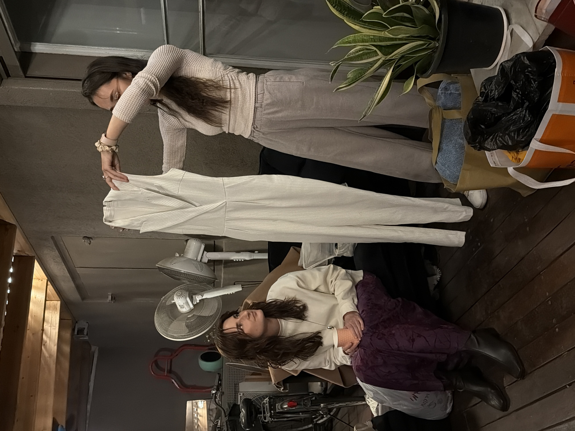 The image captures two women in an indoor setting, likely a casual living area or studio, in Tel-Aviv, Israel. The lighting suggests it's either evening or an interior space relying on artificial light.
In the upper part of the frame, a woman with long dark hair, wearing a light-colored waffle-knit top, light grey pants, and white sneakers, stands and holds up a long, white, vertically striped garment, possibly a jumpsuit or dress. She wears glasses and has a scrunchie on her left wrist, appearing to examine the item closely. To her right, a green potted plant in a black pot sits on a surface, next to a brown reusable bag containing blue fabric and an orange and white reusable bag holding a black plastic bag.
Towards the bottom, a second woman with long dark hair and glasses is seated on a wooden floor. She wears a light-colored sweatshirt, a long dark purple skirt, and black boots, her hands clasped as she looks upwards with a contemplative expression. Next to her, a white plastic bag is visible, bearing partially obscured Hebrew text including "מבצע" (Mivtza), meaning "Sale." The background features two white electric fans mounted on a wall, wooden structural beams overhead, and hints of a bicycle or machinery. The overall atmosphere is relaxed and informal.