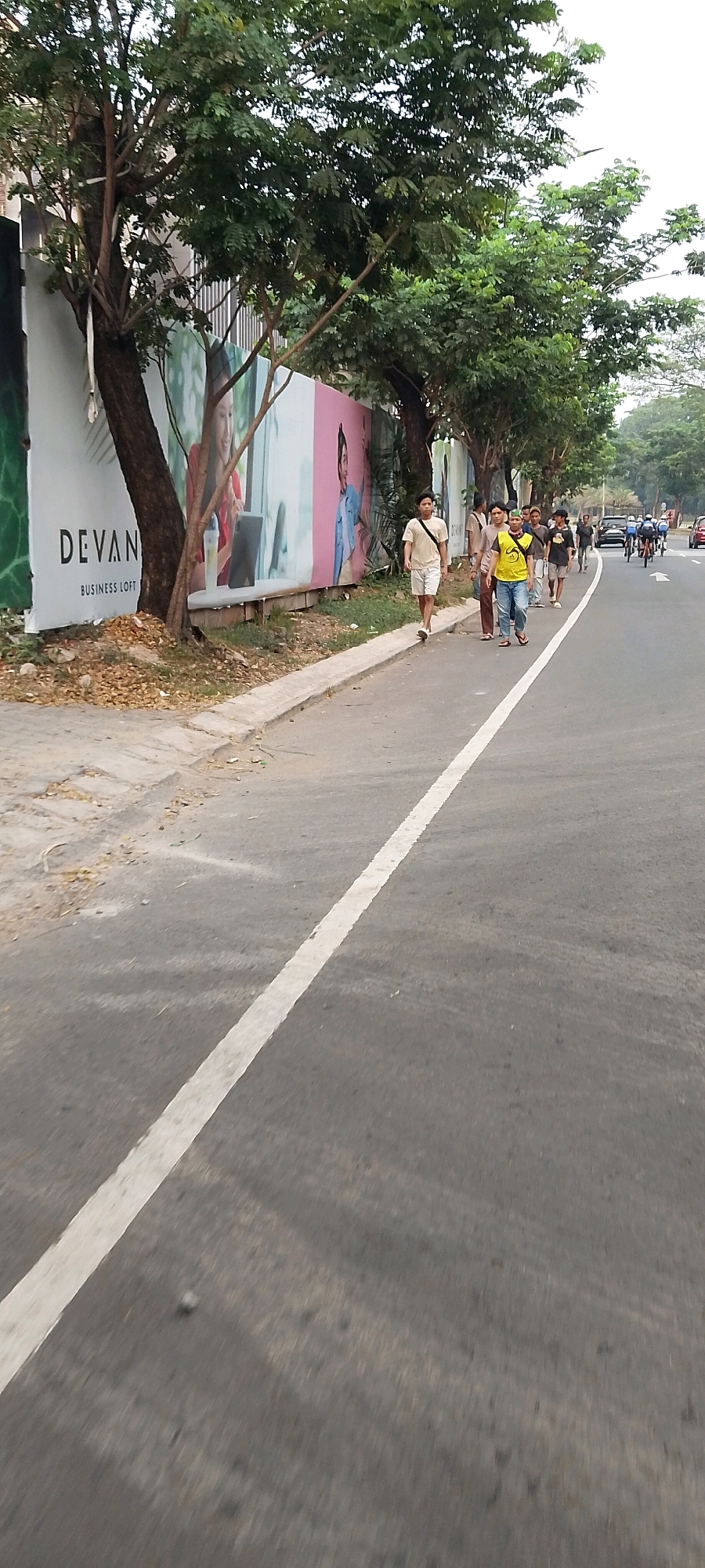 In Gunung Putri, Indonesia, a group of people walk along a paved road. The road has a white line marking the edge of the lane. On the left side of the road, a large billboard with images of people and text that reads "DEVAN BUSINESS LOFT" is visible. Trees line the billboard. The people are dressed in casual clothing. Some are walking together, while others are walking individually. The scene appears to be a normal day in a residential or commercial area.