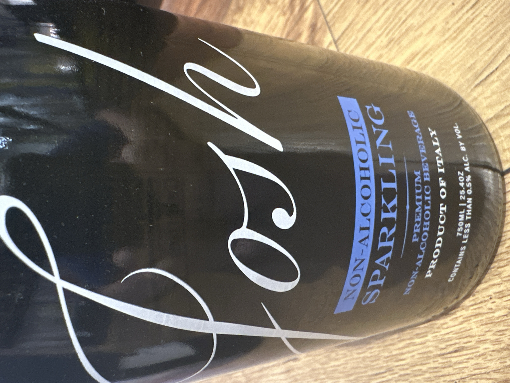 The image is a close-up, slightly angled shot of a dark bottle with white script lettering that reads "Posh". Below the "Posh" lettering, there is a blue band with the words "NON-ALCOHOLIC" in white text, followed by "SPARKLING" in larger white text. Underneath this, smaller white text reads "PREMIUM NON-ALCOHOLIC BEVERAGE" and "PRODUCT OF ITALY". The very bottom of the bottle has text stating "CONTAINS LESS THAN 0.5% ALC. BY VOL." and "750ML | 25.4OZ". The bottle is placed on a wooden surface. No specific location in Camp Hill, United States of America is visible.