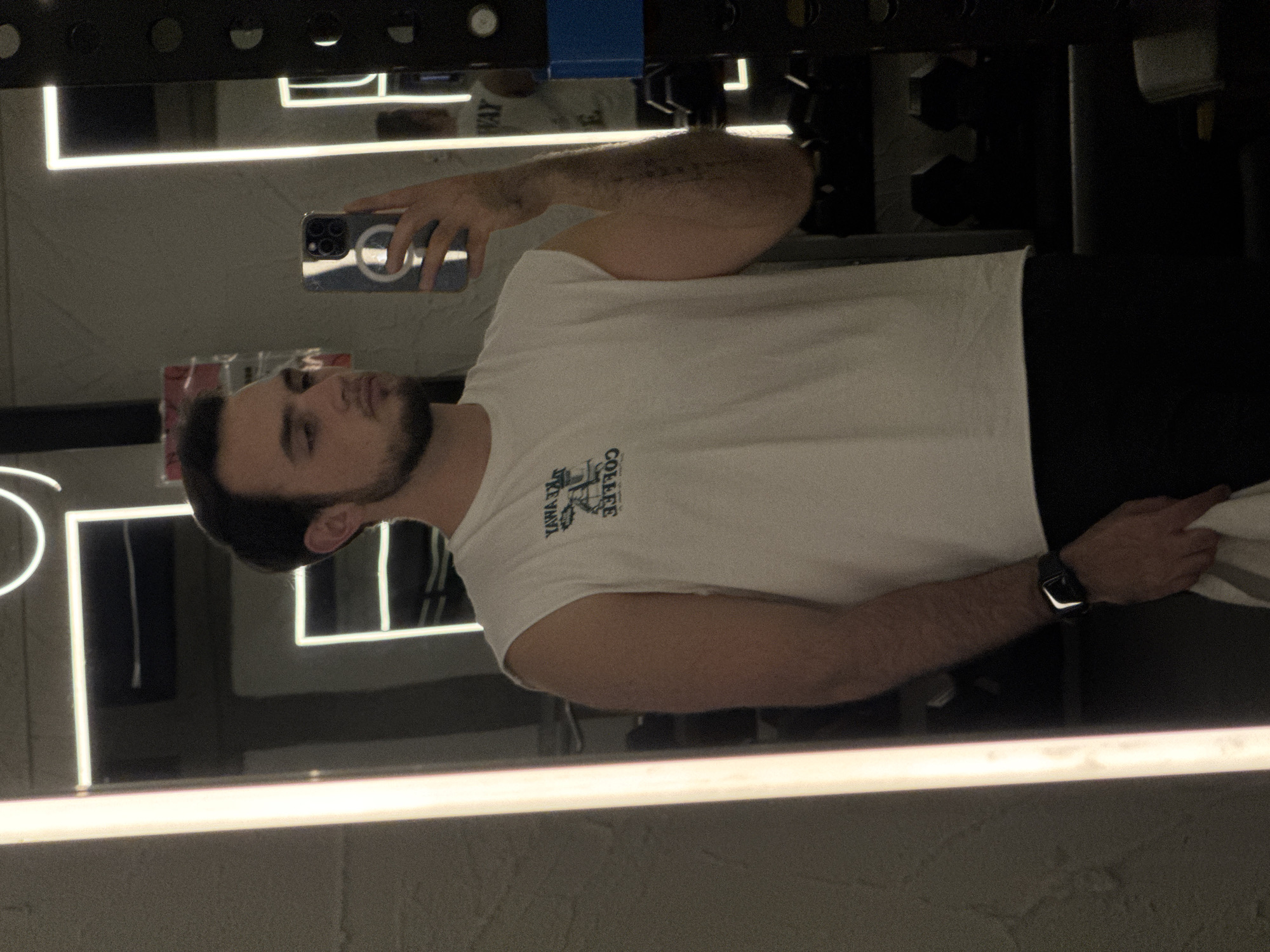 A man is taking a selfie in a mirror. He is wearing a white t-shirt and black shorts. He is holding a phone in his left hand and has a watch on his right wrist. The background appears to be a gym. It is not possible to identify the location in the city of Puebla, Mexico with the information provided.