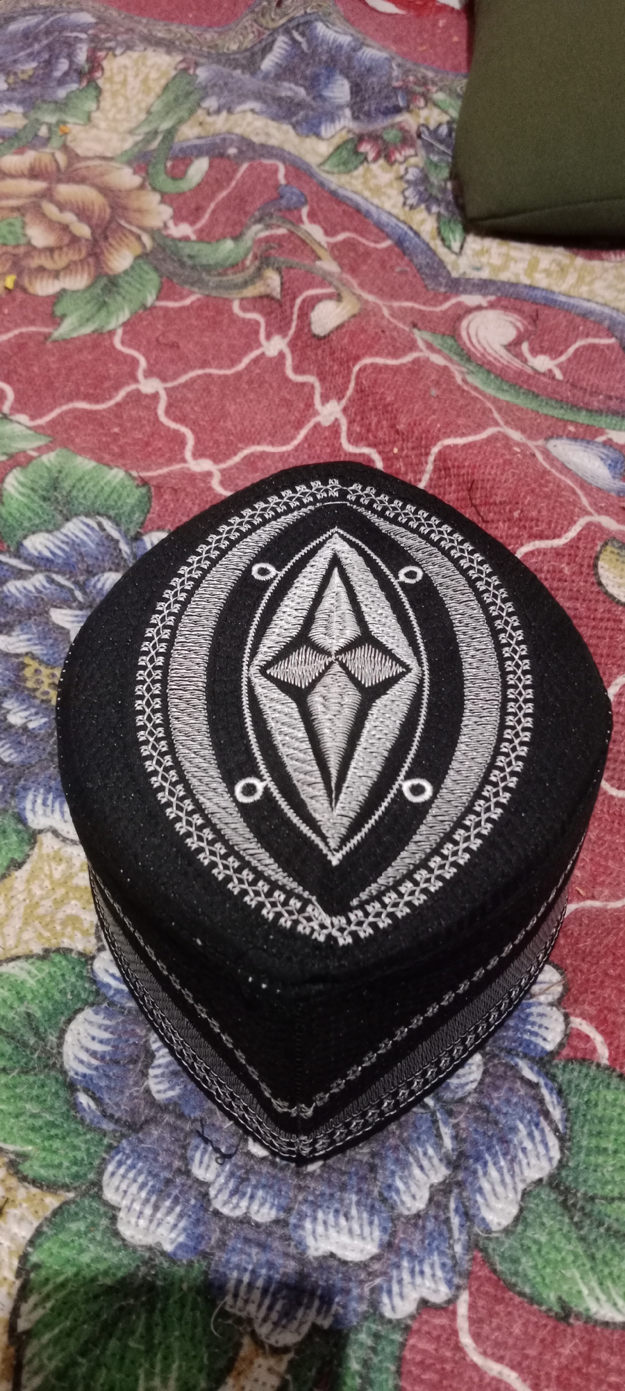 A black kufi with intricate silver embroidery is placed on a fabric surface. The kufi is cylindrical with a flattened top, and its embroidery forms a diamond shape surrounded by a circular pattern. The fabric beneath it features a floral design with red, blue, and green hues. The background is out of focus, but appears to be another patterned fabric. The lighting suggests an indoor setting. There are no people visible in the image. The location in Soko, Indonesia, is not discernible from the visual information provided.