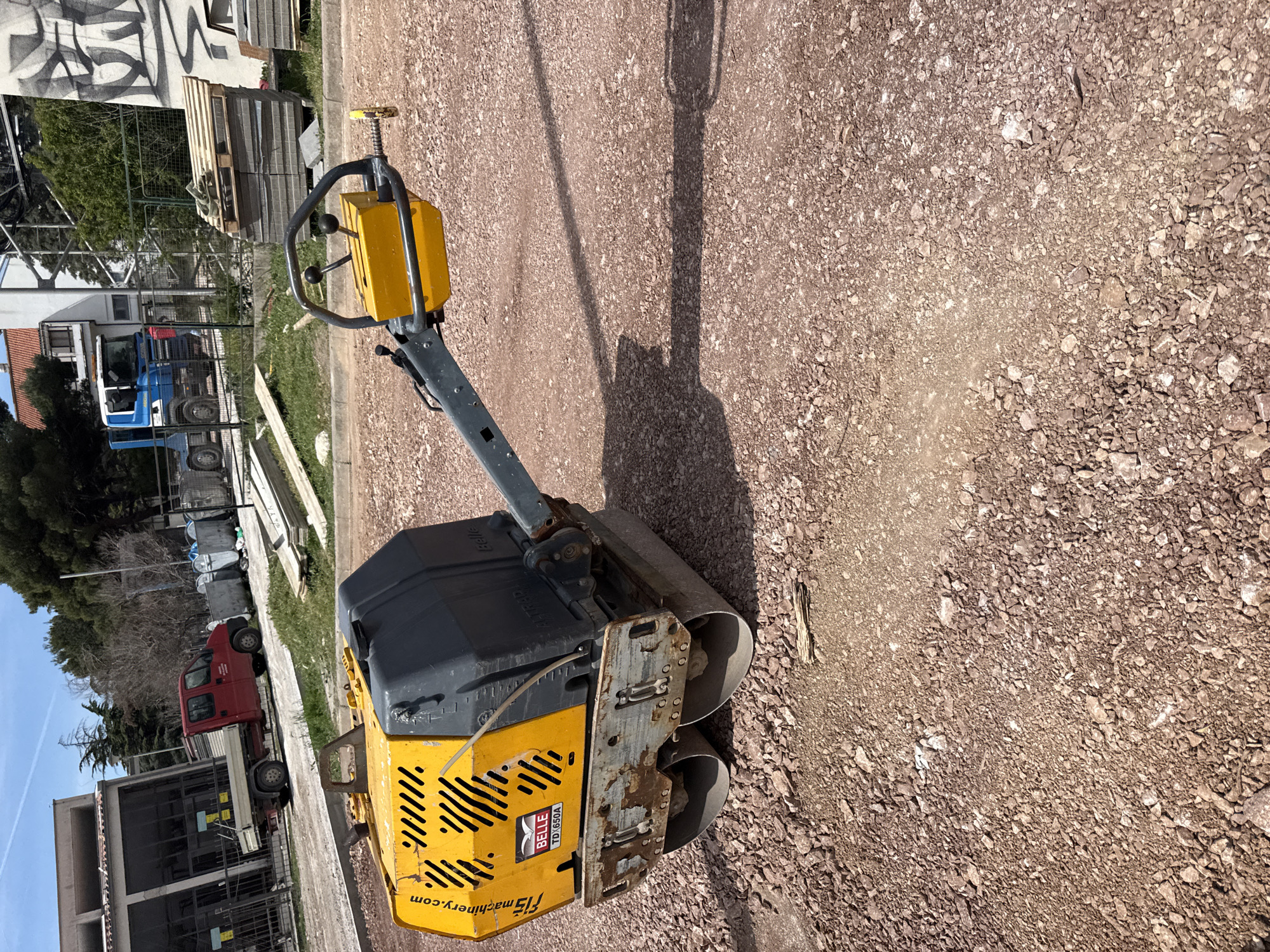 The image shows a Belle TD1.650A roller compactor on a gravel surface. In the background, there are buildings, parked vehicles, and construction materials like pallets and wooden planks. The location appears to be a construction or work site in an urban area.