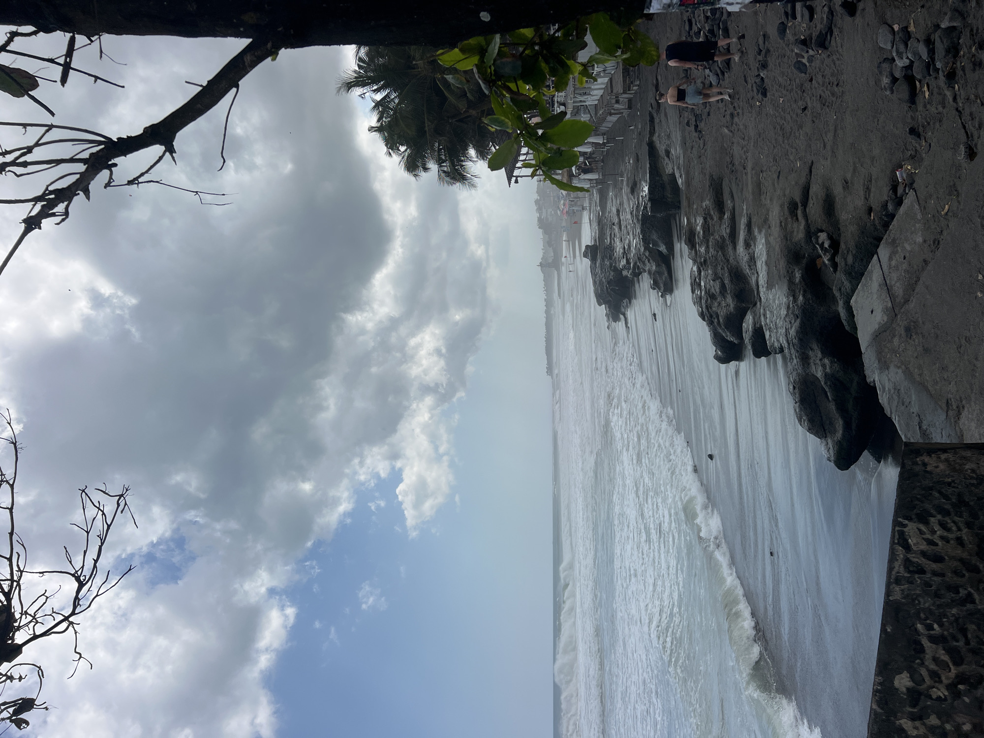 The image captures a view of the coastline in North Kuta, Indonesia, under a cloudy sky. The ocean on the left features moderate waves crashing onto a dark sand beach. The beach is bordered by dark, rocky formations and a concrete walkway. Further along the coastline, in the distance, several buildings with thatched roofs and umbrellas are visible, suggesting a beachside establishment. A few palm trees are scattered along the beach. Two individuals, a woman in a dark top and shorts and a man in a dark t-shirt and shorts, are walking away from the camera along the beach. The foreground on the right is dominated by the dark trunk of a tree and some green leaves, partially obscuring the view of the beach and the ocean.