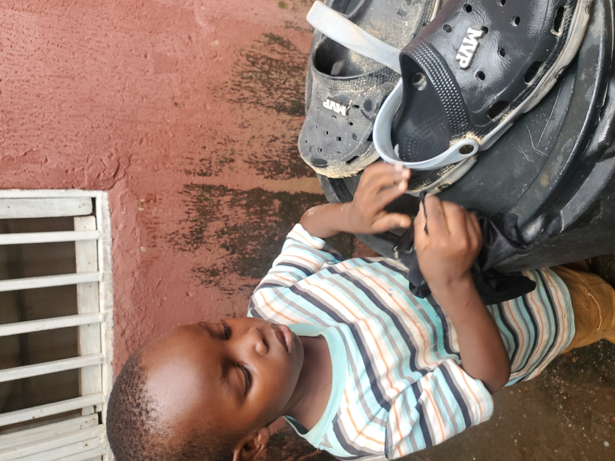 A young boy, likely under ten years of age, is captured in a medium shot, looking down with a focused expression as he cleans a pair of black Croc-style sandals. He wears a horizontally striped t-shirt with colors of light blue, white, orange, and dark blue, and tan pants. The sandals, resting on what appears to be a black plastic container, are somewhat dirty. The background consists of a reddish-brown textured wall, typical of the architecture in many parts of Jos, Nigeria, and a section of a white-barred window with a concrete sill. The ground is wet and appears to be dirt or pavement, suggesting a recent rain or a damp environment. The overall scene evokes a sense of everyday life and domestic activity in an urban Nigerian setting.