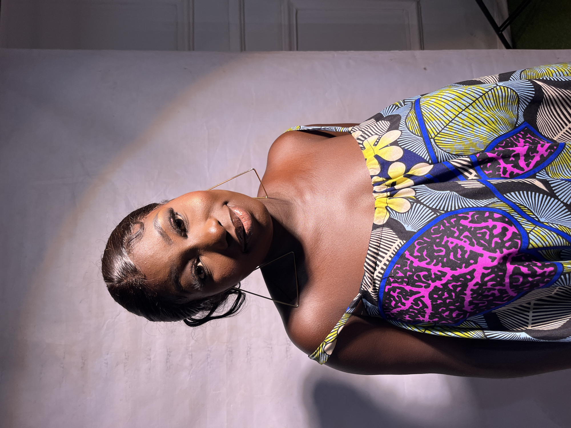 A medium shot shows a young Black woman with dark skin and dark hair pulled back, wearing large, gold, triangular hoop earrings and a colorful strapless dress. The dress has a vibrant African print with bold patterns of pink, blue, yellow, and black. She is looking at the camera with a slight smile, her head tilted. Her makeup includes dark eyeliner and mascara, and a shimmery lip gloss. The background is a plain, textured light-colored wall, with a subtle diagonal light beam or shadow cast across it from the upper left to the lower right. There is also a soft shadow on the lower left of the image. The lighting suggests an indoor studio or a well-lit indoor space. There are no discernible elements in the image to indicate a specific location within Jos, Nigeria.