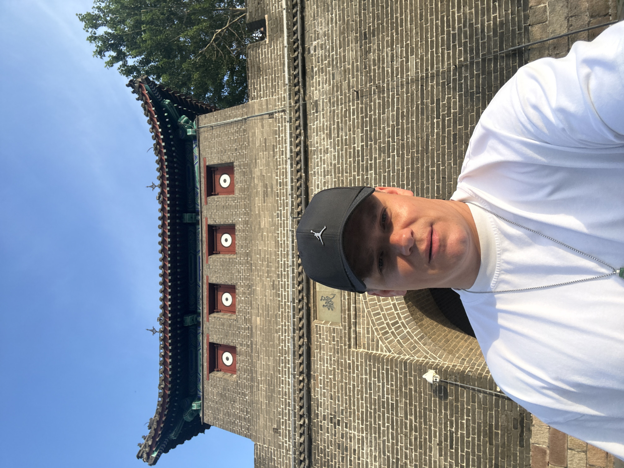 A man wearing a white t-shirt and a black baseball cap is taking a selfie in front of a stone gate structure. The man's face is visible from the nose up, and he is looking directly at the camera. The gate structure is made of grey bricks and has a traditional Chinese architectural style with a curved roof decorated in red and green. Above the archway of the gate, there is a stone carving with Chinese characters, and three square windows with red frames and white circles are visible on the upper part of the gate. A tree is partially visible on the right side of the image. The sky is clear and blue. The location is identified as the Shanhaiguan Great Wall in Qinhuangdao, China.