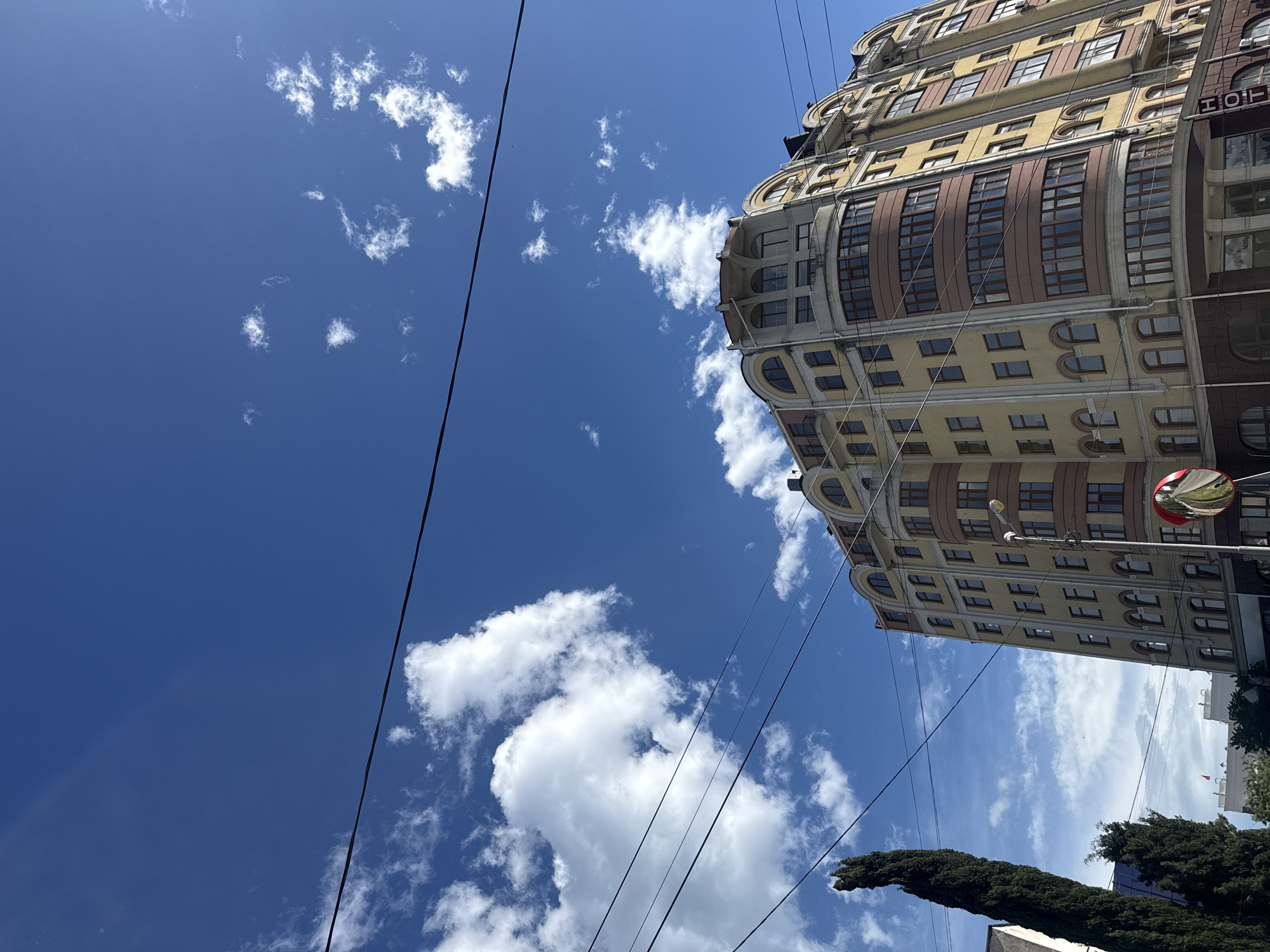 The image shows a tall building in Unknown, Unknown, viewed from a low angle. The building is multi-story, with a curved facade on one section, and features a mix of brown and yellow colors with numerous windows. The sky is visible, filled with fluffy white clouds against a deep blue background. Electrical wires crisscross the view, cutting through the sky and near the building. Trees are partially visible at the bottom of the image.