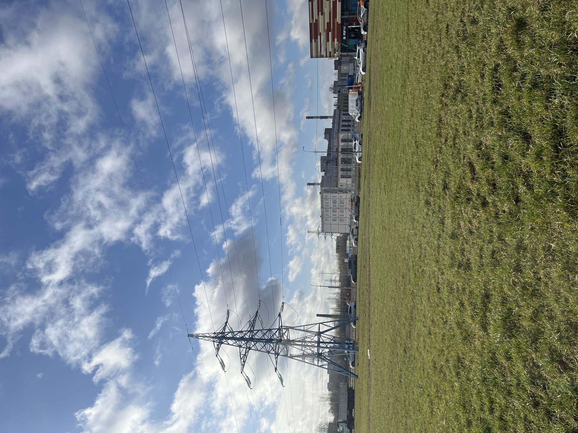 The image shows a green grassy field in what appears to be an urban environment in Saint Petersburg, Russia. An electrical transmission tower, painted blue, dominates the left side of the frame, with power lines stretching into the distance. In the background, buildings of various types are visible, including what seems to be an industrial structure with smokestacks. There are parked cars and what looks like a modern, multi-colored building in the background. The sky is blue with scattered white clouds.