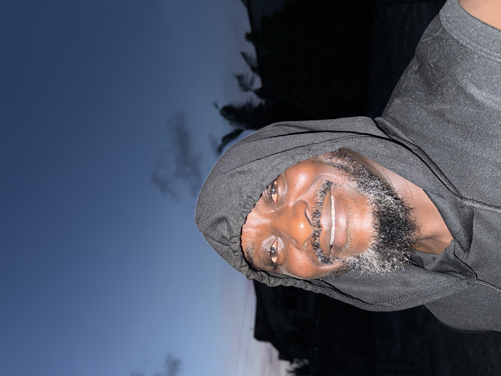 A medium-shot, portrait-style photograph shows a Black male wearing a dark hooded sweatshirt. His hood is up. He faces the camera and offers a slight smile. He has a mustache and beard that appear to be salt and pepper. The backdrop shows a twilight sky with faint colors of the setting sun and palm trees. The location is likely Ado, Nigeria, given the request.