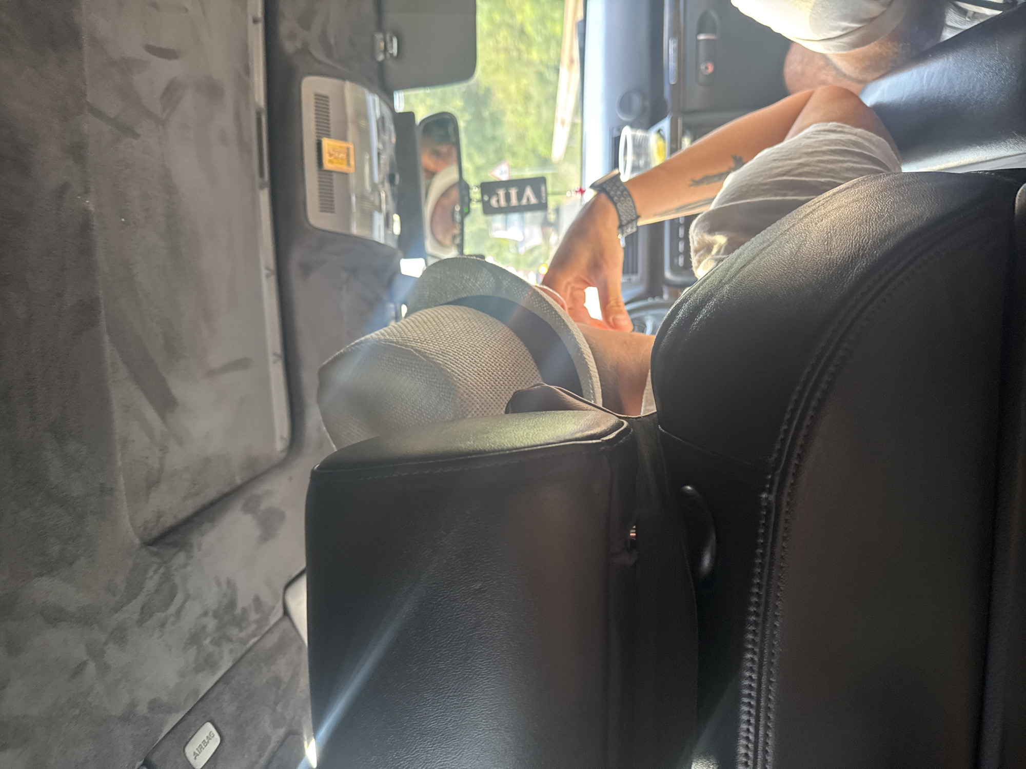 The image shows the interior of a car, viewed from the back seat. A person wearing a hat is seated in the front passenger seat, facing away from the camera. The person's right arm is visible, resting on the dashboard and showing a tattoo. A cup with a yellow liquid is also on the dashboard. A "VIP" air freshener hangs from the rearview mirror, which reflects a person's face. The car's ceiling has a textured gray upholstery, with an "AIRBAG" label visible. The exterior of the car, seen through the windshield, suggests a daytime scene with greenery, indicating a likely location outside a city, possibly on a road leading to or from Tsarevo, Bulgaria. The overall impression is of a relaxed journey during daylight.
