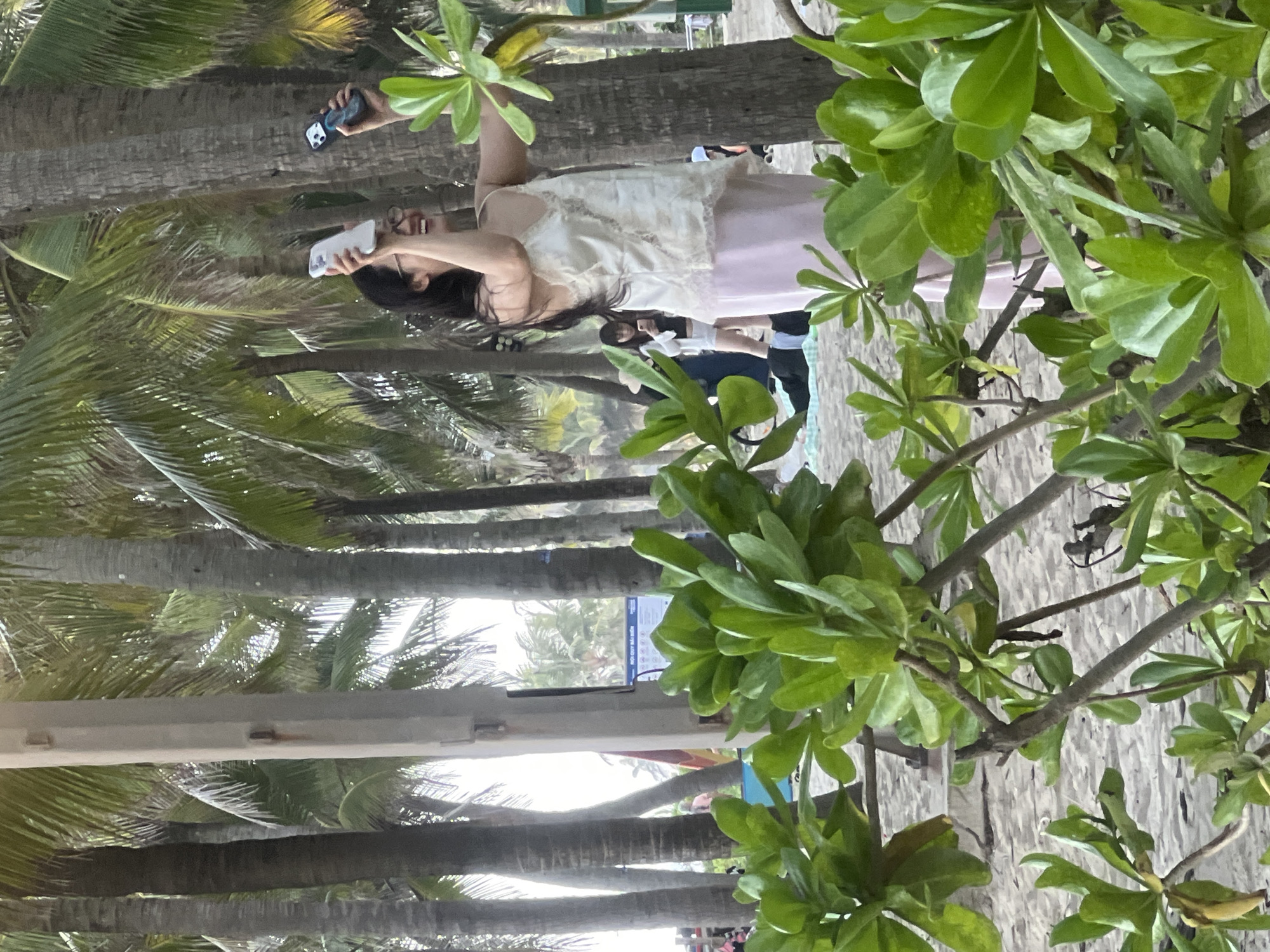 The image depicts a scene at a beach in Đà Nẵng, Vietnam, characterized by the presence of palm trees and sandy ground. A woman in the foreground, wearing a light-colored top and a pink skirt, is holding up a white smartphone, seemingly taking a selfie. Her expression is one of happiness and amusement. Behind her, other individuals are visible, including someone holding a dark-colored smartphone, and further back, several people are gathered. The lighting suggests it is daytime, possibly late afternoon, with sunlight filtering through the palm fronds. Green foliage from a tree in the foreground partially obstructs the view of the background. A blue sign with text is partially visible in the mid-ground. The overall atmosphere is relaxed and vacation-like.