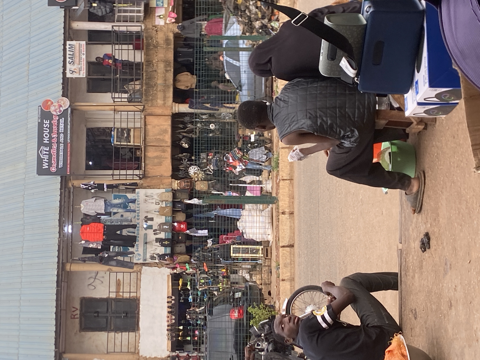 The image shows an urban scene in Bukuru, Nigeria. There are two individuals: one seated and working on an object, and another lying down and looking up. A building is visible with businesses such as "White House Cosmetics & Jewelry" and "Salim Collections" with merchandise displayed outside. There is also a sign for "SD Sirobbex Collection" visible. There is a collection of bags and various items on a makeshift table beside the seated man. A motorcycle is parked nearby the man lying down. The street is paved.