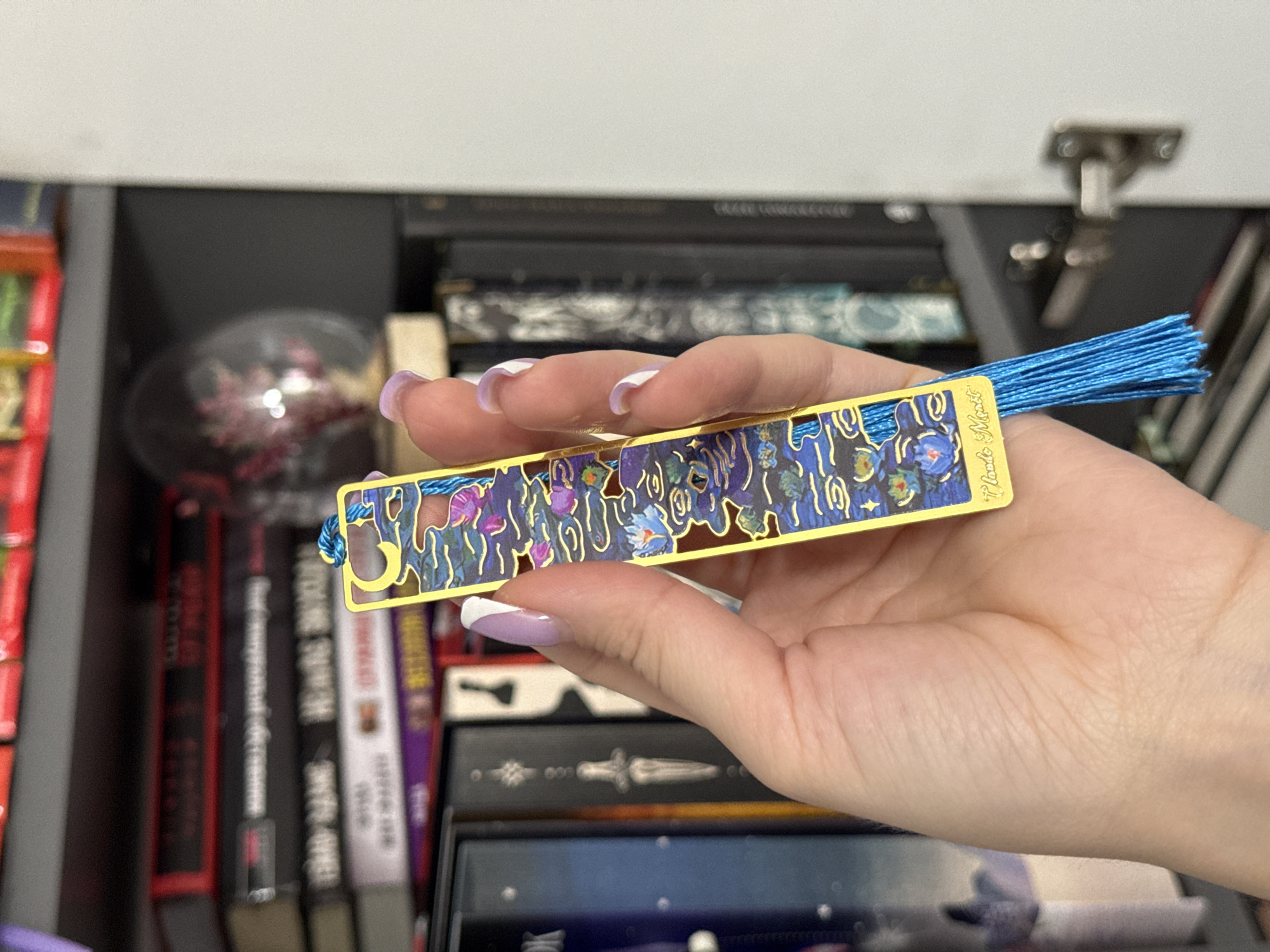 A close-up shot shows a person's hand holding a gold bookmark. The bookmark features a decorative cutout design with a floral motif and a blue tassel. The background is blurred, revealing shelves filled with books, suggesting a library or bookstore setting. The lighting appears to be indoor, with a soft, diffused light source. The hand has a light skin tone and neatly manicured nails painted with a light purple polish and white tips. The image is cropped to focus on the hand and bookmark, with the background details softly out of focus. The overall impression is one of a quiet, organized space, possibly a personal collection of books or a retail environment. There is no clear indication of the time of day or weather conditions. The location context of Łódź, Poland is not visually evident in the image itself.