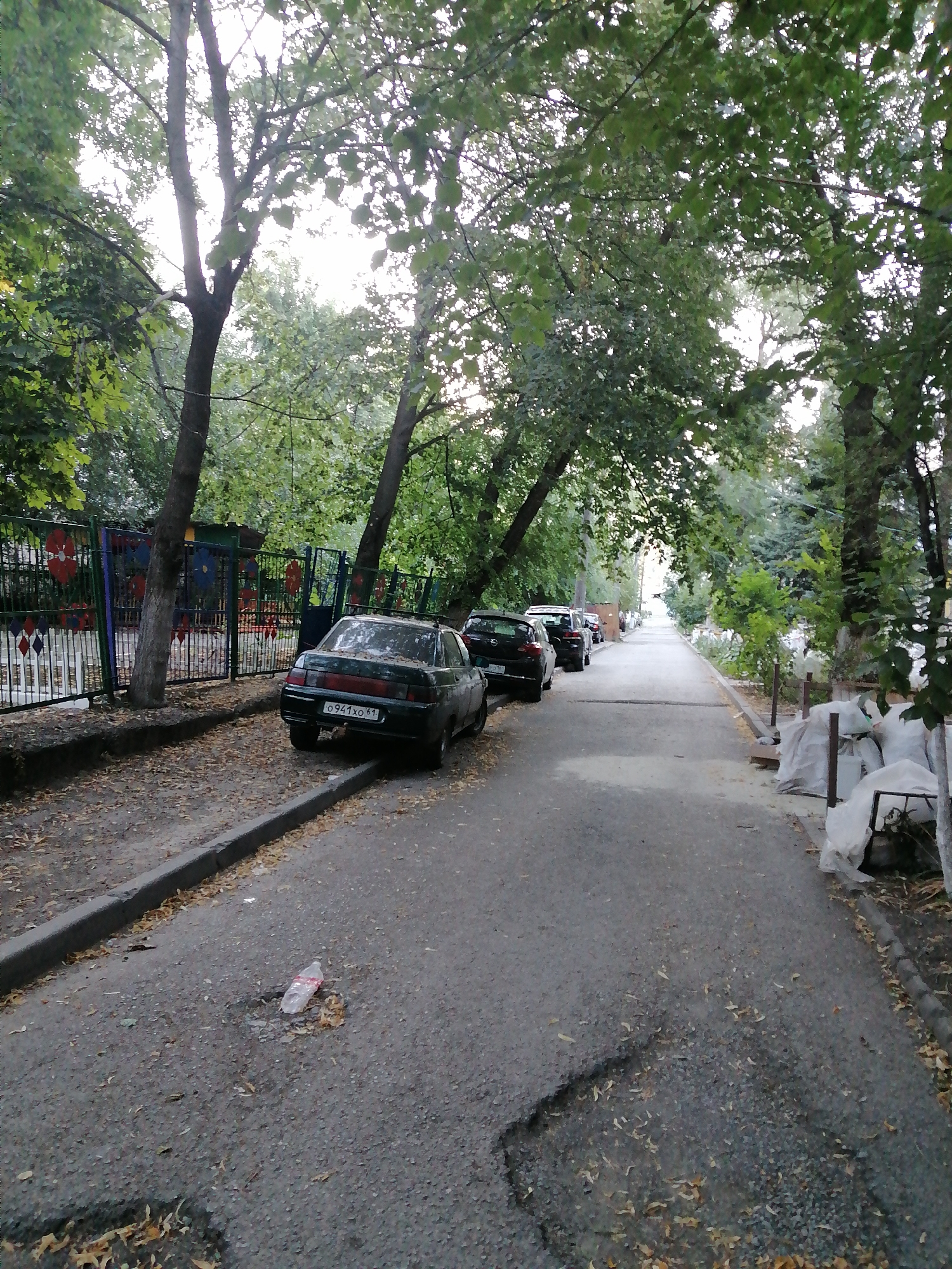 The image depicts a street in Rostov-on-Don, Russia, lined with mature trees casting shadows on the asphalt. A black sedan with a license plate reading "O941XO 61" is parked on the right side of the road, facing away from the camera. The car appears to be dusty and covered with fallen leaves, suggesting it has been parked for some time. To the left of the car, a low fence with colorful floral decorations separates the street from a green area. Further down the street, more cars are parked along the roadside, and the path appears to be sparsely populated. The overall impression is one of a quiet, residential street in a city with a significant amount of greenery.