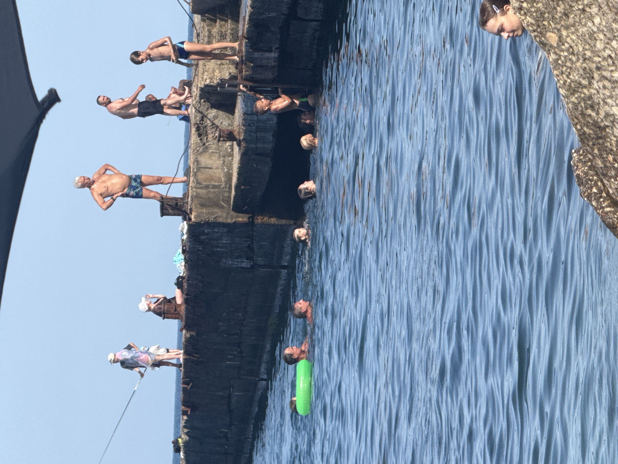The image depicts a sunny day at a concrete pier in Partenit, Ukraine, with people enjoying the sea. Several individuals are visible on the pier, including a man in floral swim trunks with his hands on his hips, another man in black swim trunks looking down, and a boy in blue swim trunks. Further along the pier, a person is fishing with a long rod, and a woman in a black top and white cap is seated, drinking from a bottle. In the water, a group of people are swimming, with some visible heads and a green inflatable ring. A young girl is also seen in the water near the foreground. The environment consists of a clear blue sky, calm blue sea, and a rough, weathered concrete pier.