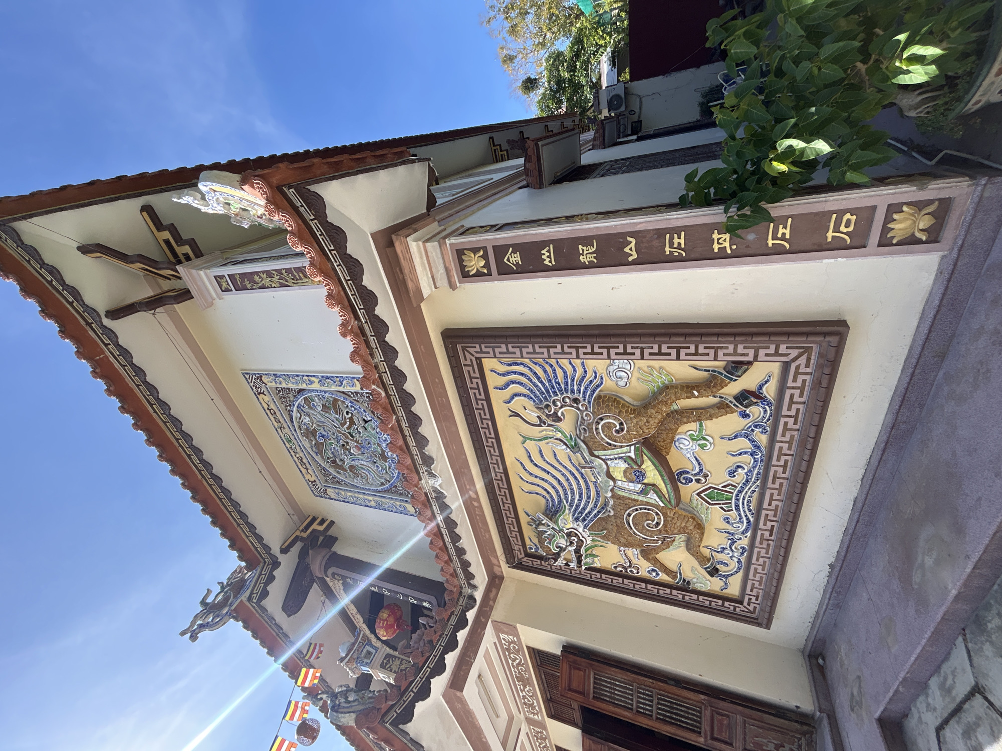 The image shows the exterior architectural details of a building in Tay Nha Trang Ward, Vietnam. The building features ornate tiled artwork on the exterior walls and under the eaves. One prominent tiled panel depicts two mythical creatures. The walls are painted in muted tones with dark brown trim. Chinese characters are inscribed in gold on a dark band. The sky is clear blue. The photograph is taken from a low angle, looking upwards.