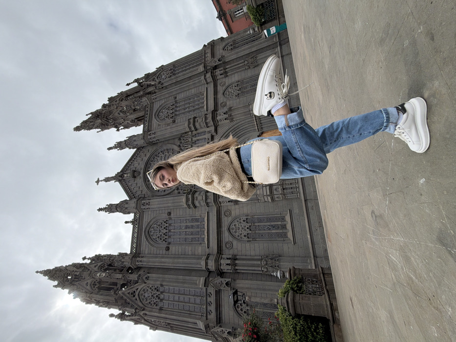 An outdoor, eye-level full-shot captures a young female posing in front of the Parroquia de San Juan Bautista in Arucas, Gran Canaria, Spain. The female, centered in the frame, has long blonde hair and wears a beige teddy-style bomber jacket, light blue baggy jeans, white platform sneakers, and white rectangular sunglasses atop her head. She carries a light-colored "VALENTINO" cross-body bag. Her left leg is raised, bent at the knee, with her foot positioned behind her right calf. She looks directly at the camera over her right shoulder, her lips slightly pursed. The gothic revival church, constructed from dark grey stone, dominates the background, featuring multiple spires, intricate tracery windows, and detailed ornamentation. The sky is overcast with a bright spot visible behind the church's left spire. The ground beneath the subject is a light grey paved plaza. To the left, dark green shrubbery and a black street lamp are partially visible. A reddish-orange building roof is visible on the far right.