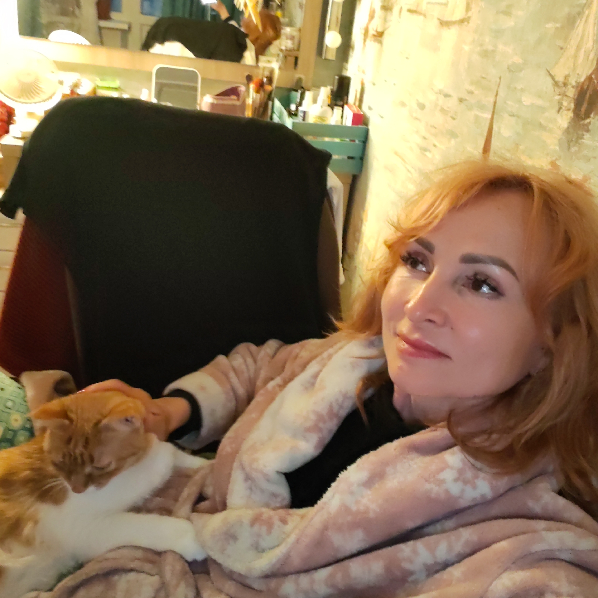 A woman with light auburn, wavy hair is shown from the chest up, in a domestic indoor setting. She is wearing a pink and white patterned robe over a black top. She is looking upward and to her right, and resting her hand on an orange tabby cat nestled in her lap.
Behind her is a black-covered chair. In the background, a vanity area with a mirror, various makeup items, a small white fan, and a light green container holding cosmetics are visible. The wall is pale yellow with textured patterns.
There is no information to verify that this image was taken in Chaikovsky, Russia.