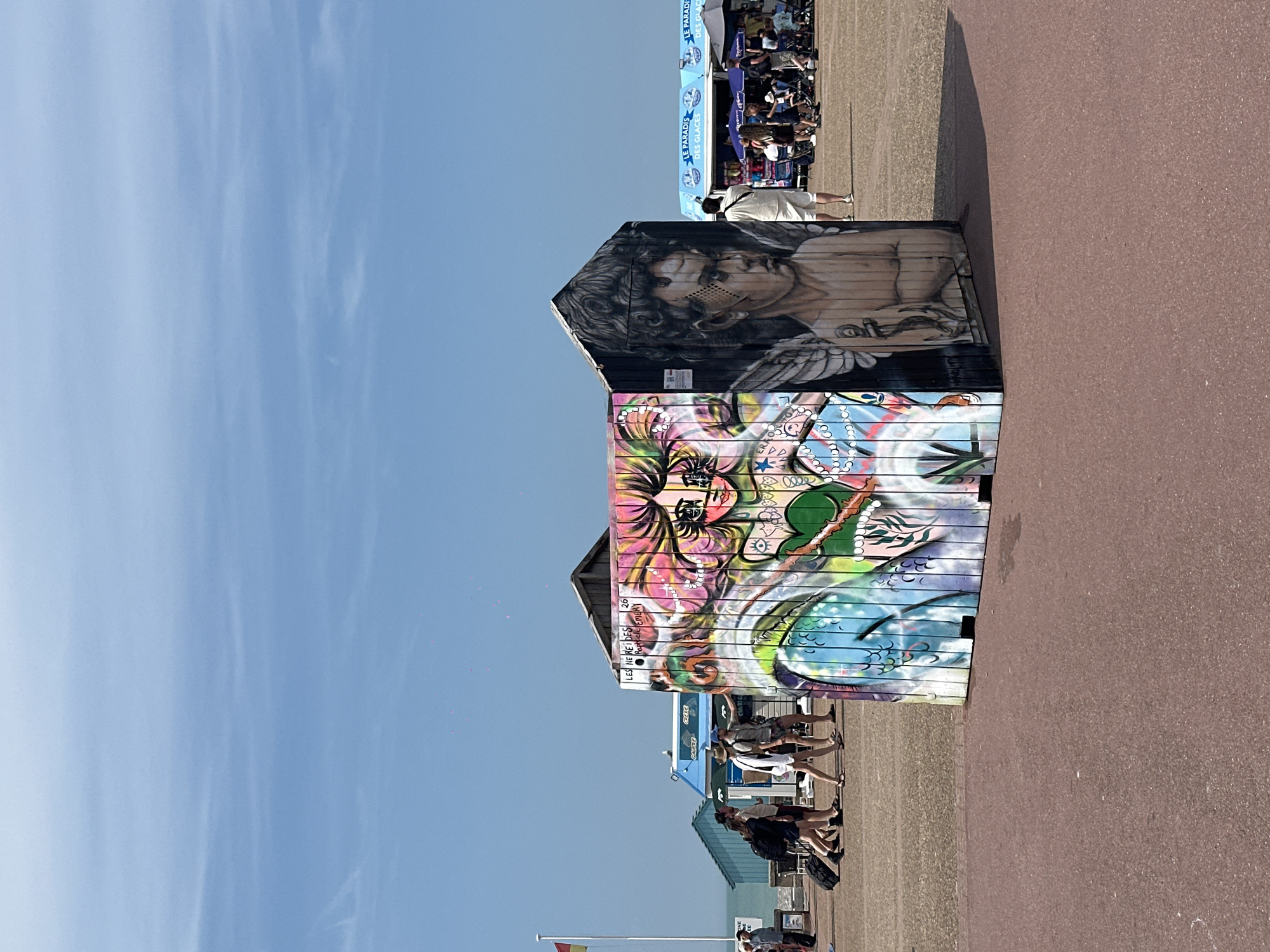 This image depicts a vibrant, graffiti-covered structure situated on a paved promenade in Dieppe, France. The structure, resembling a small house or shed, is adorned with colorful graffiti featuring a woman's face with flowing pink hair on one side, and a black and white portrait of a young person with wings and an anchor tattoo on the other. The promenade is populated by several individuals walking and standing. In the background, there are beach huts, ice cream vendors, and more people enjoying the outdoor setting. The sky is a clear, bright blue with a few faint wispy clouds. The overall scene suggests a lively, public space, likely a beach or boardwalk area in Dieppe, on a sunny day.