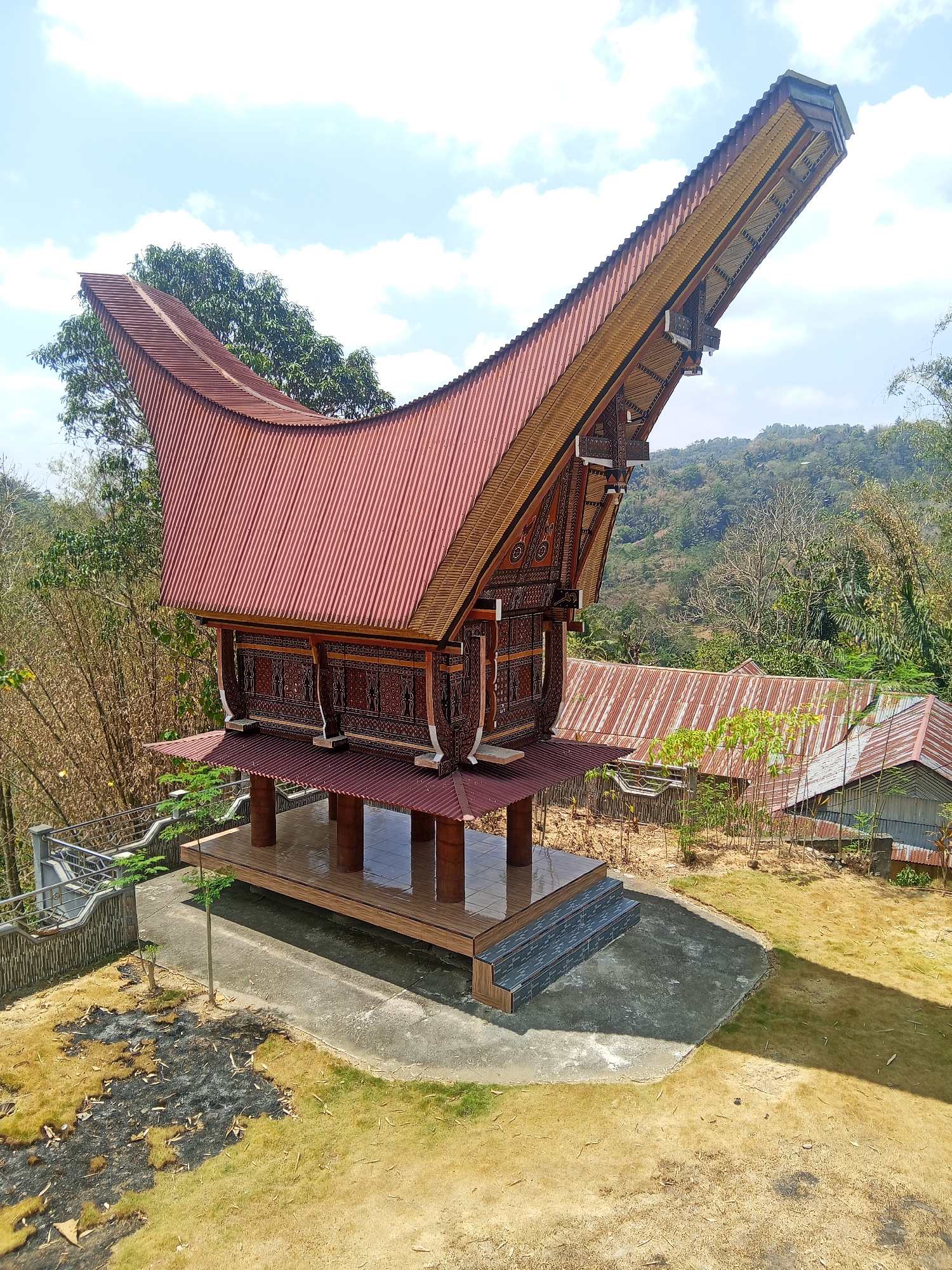 The image shows a traditional Tongkonan house in Batu Papan, Indonesia. The Tongkonan is a traditional house of the Toraja people, and it is characterized by its distinctive boat-shaped roof. In this image, the Tongkonan is elevated on stilts and features intricate carvings on its wooden facade. The roof is made of red corrugated metal. The surrounding environment is lush and green, with trees and hills visible in the background. There are also some other buildings with corrugated metal roofs in the distance, suggesting a rural or semi-rural setting. The sky is blue with scattered clouds, indicating a sunny day. The ground in front of the Tongkonan is a mix of concrete and dry grass, with some patches of burnt vegetation. There are no people visible in the image.