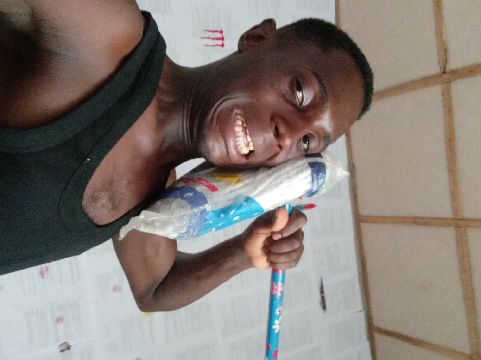 A close-up shot depicts a man, likely an adult male, holding a wrapped item against his cheek. He wears a black tank top. The man's skin tone is dark, and he displays a wide grin, revealing his teeth. The wrapped item, possibly a pillow or cushion, is encased in clear plastic with blue and white decorative elements. The man is holding a blue pole with white floral designs on it. The background is a wall covered in a white material. To the right of the man is a wall with beige and brown wooden beams. The location, Akwete, Nigeria, is not directly identifiable from the visual elements in the photograph. No interactions are visible in the image.